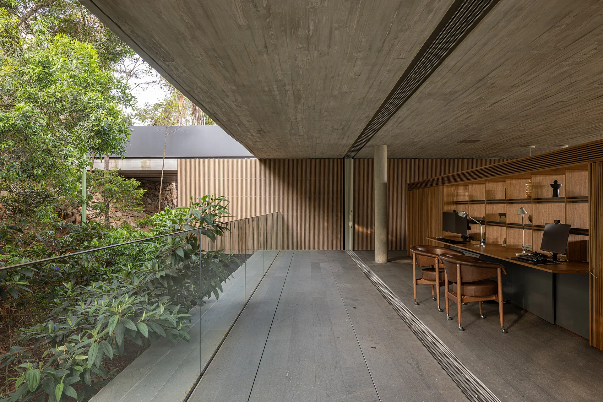 A modern home office featuring a built-in wooden desk under a concrete ceiling, opening directly onto an outdoor terrace with a glass railing.