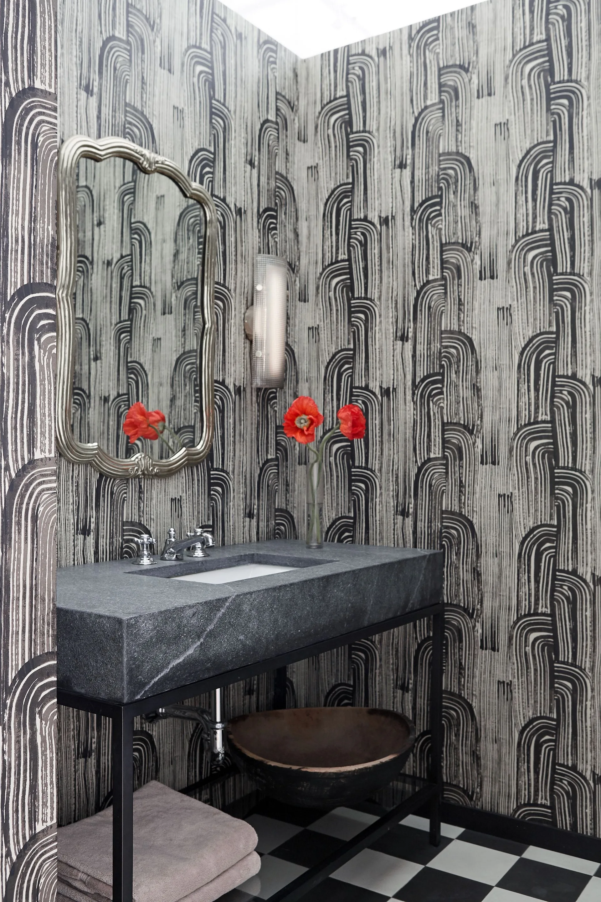 A powder room features a dark stone vanity, an ornate silver mirror, and bold black and white arched wallpaper above checkered floor tiles.