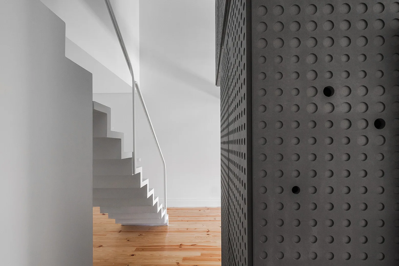 A minimalist white floating staircase ascends past a dark charcoal perforated metal cabinet on light wood flooring.