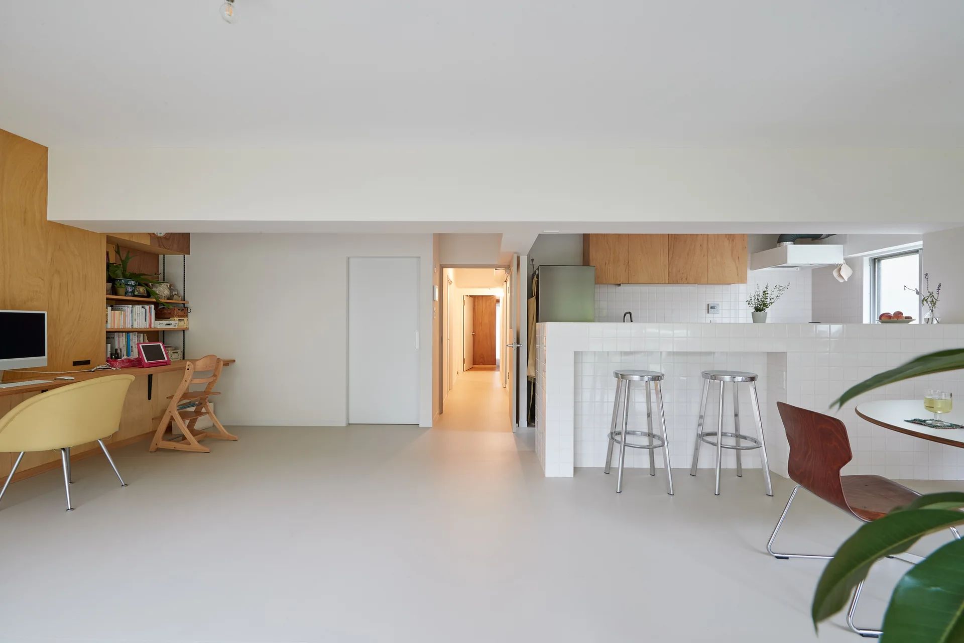 A minimalist open-plan room features a glossy white-tiled kitchen island with metal stools alongside a built-in light wood study desk on a white wall.