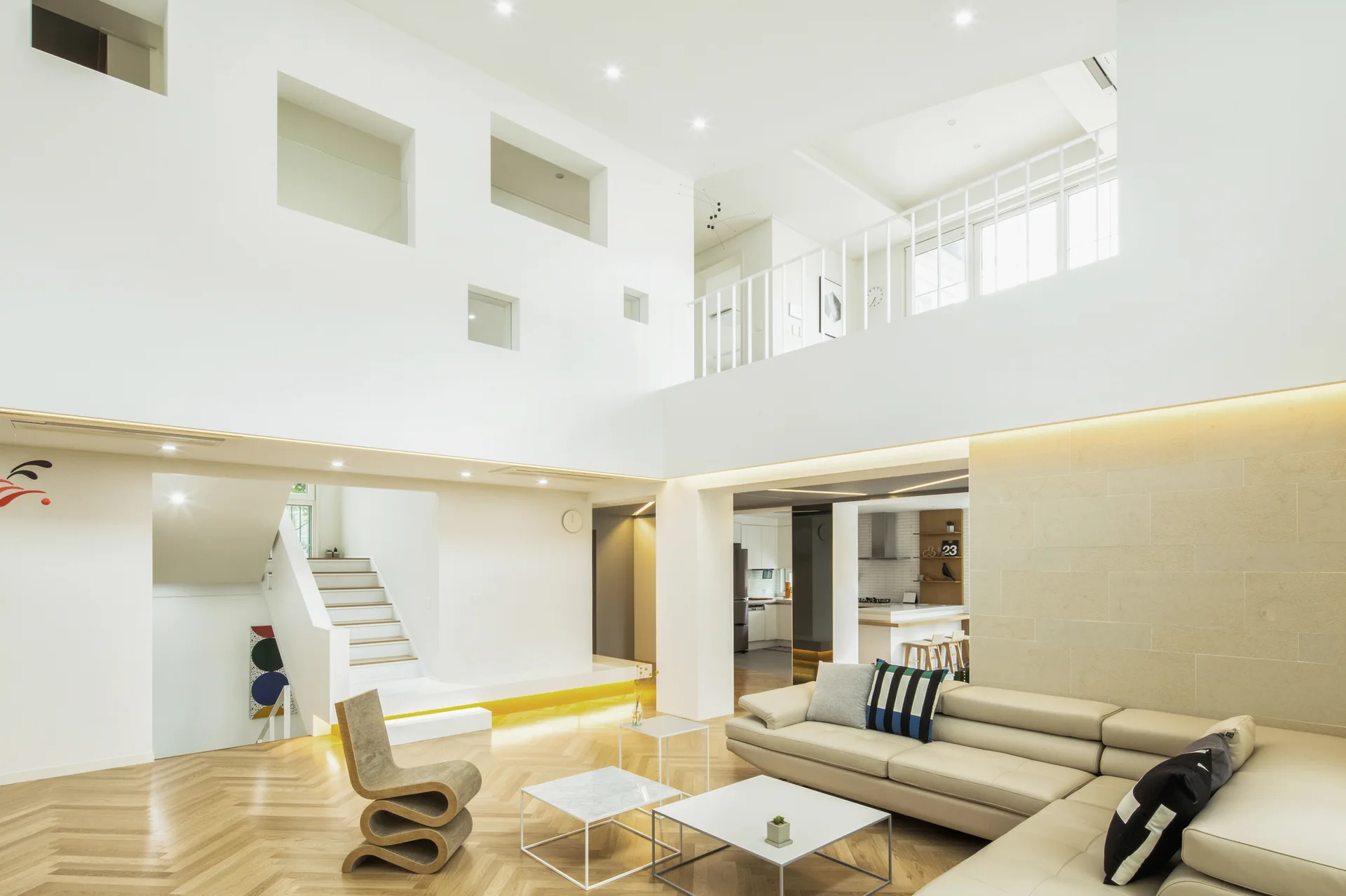 A double height white living room with herringbone wood floors, a cream sectional sofa, a sculptural zigzag chair, and square interior wall cutouts.