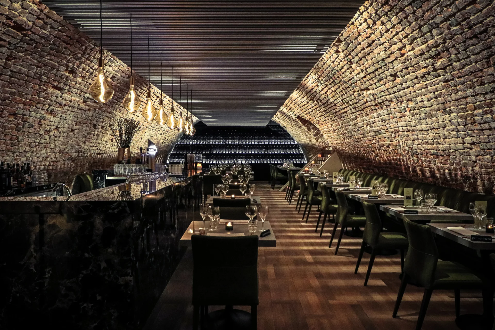 Restaurant dining room featuring a curved brick barrel-vaulted ceiling, a polished dark bar counter, and rows of tables with olive green chairs.