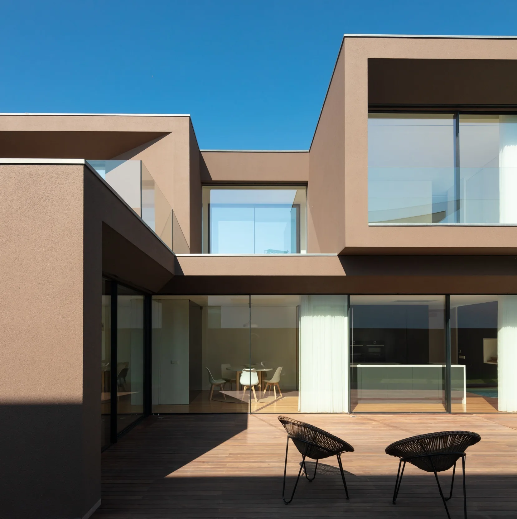 A modern two-story brown stucco house features large glass sliding doors that open onto a wooden deck with two black wire chairs.