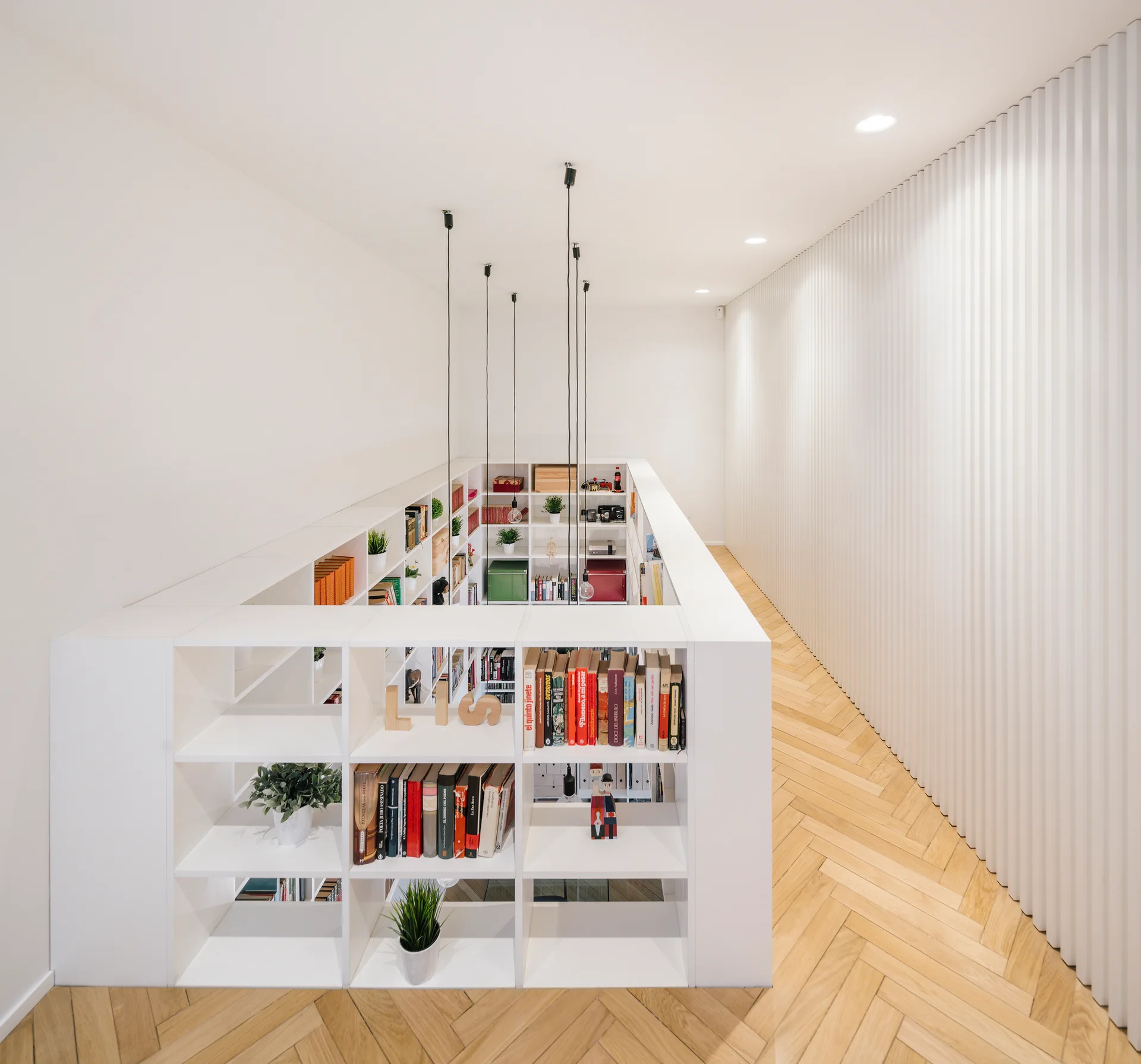 A white hallway landing features a central built-in bookshelf surrounding a stairwell, herringbone wood flooring, and a fluted wall.