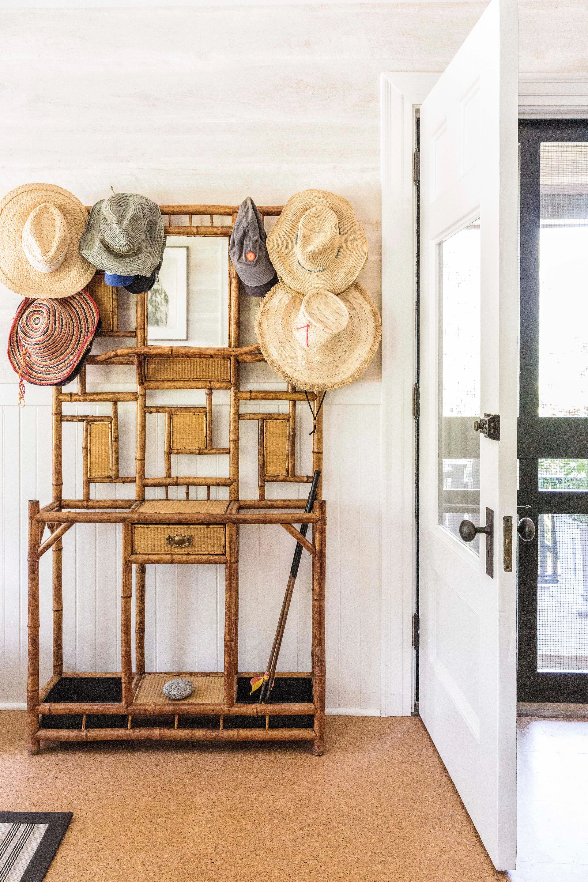 A vintage bamboo hall tree holding several straw hats stands against a white paneled wall near an open door on cork flooring.