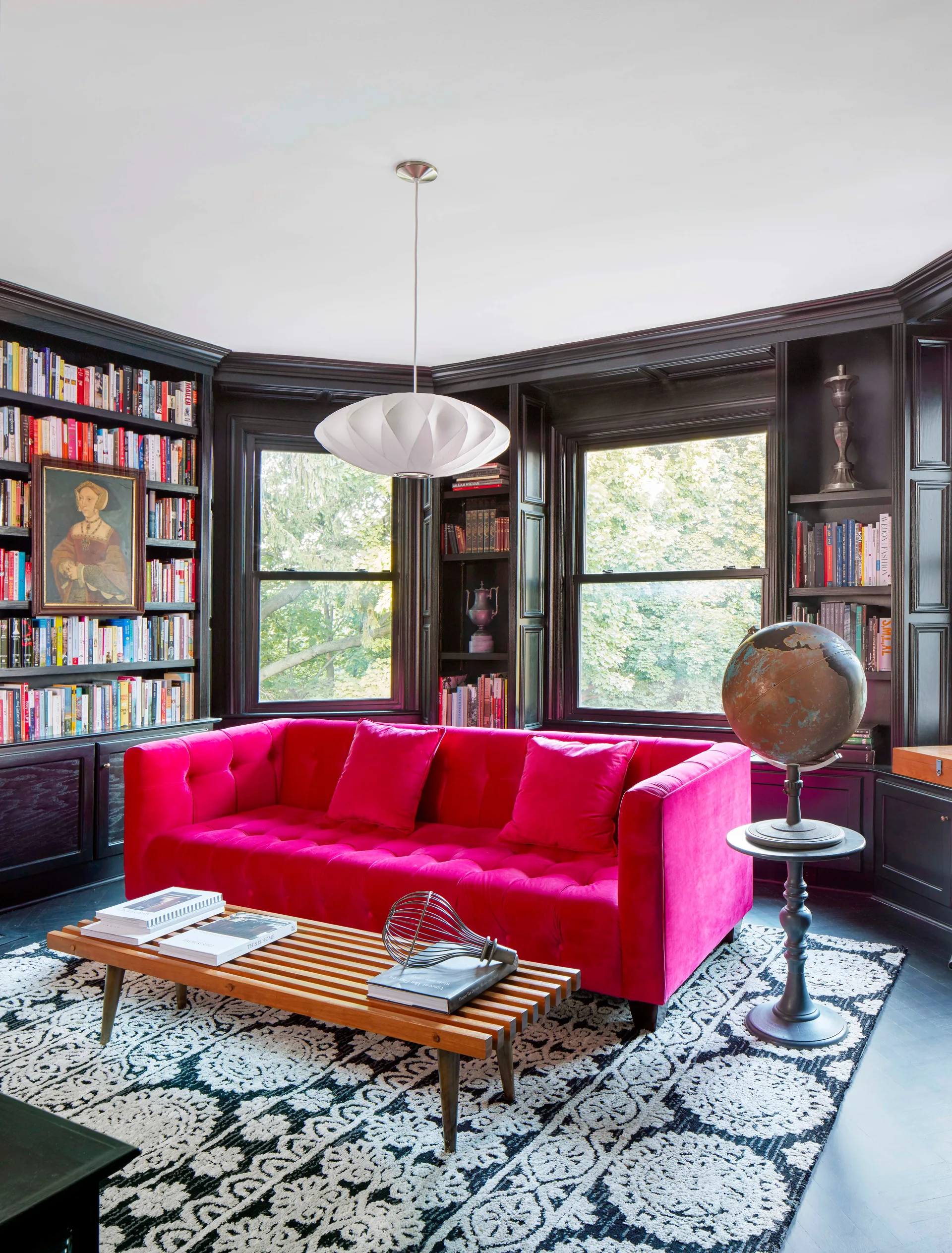 A bright pink velvet sofa sits in front of black built-in bookshelves and a bay window, with a slatted wood coffee table in the foreground.