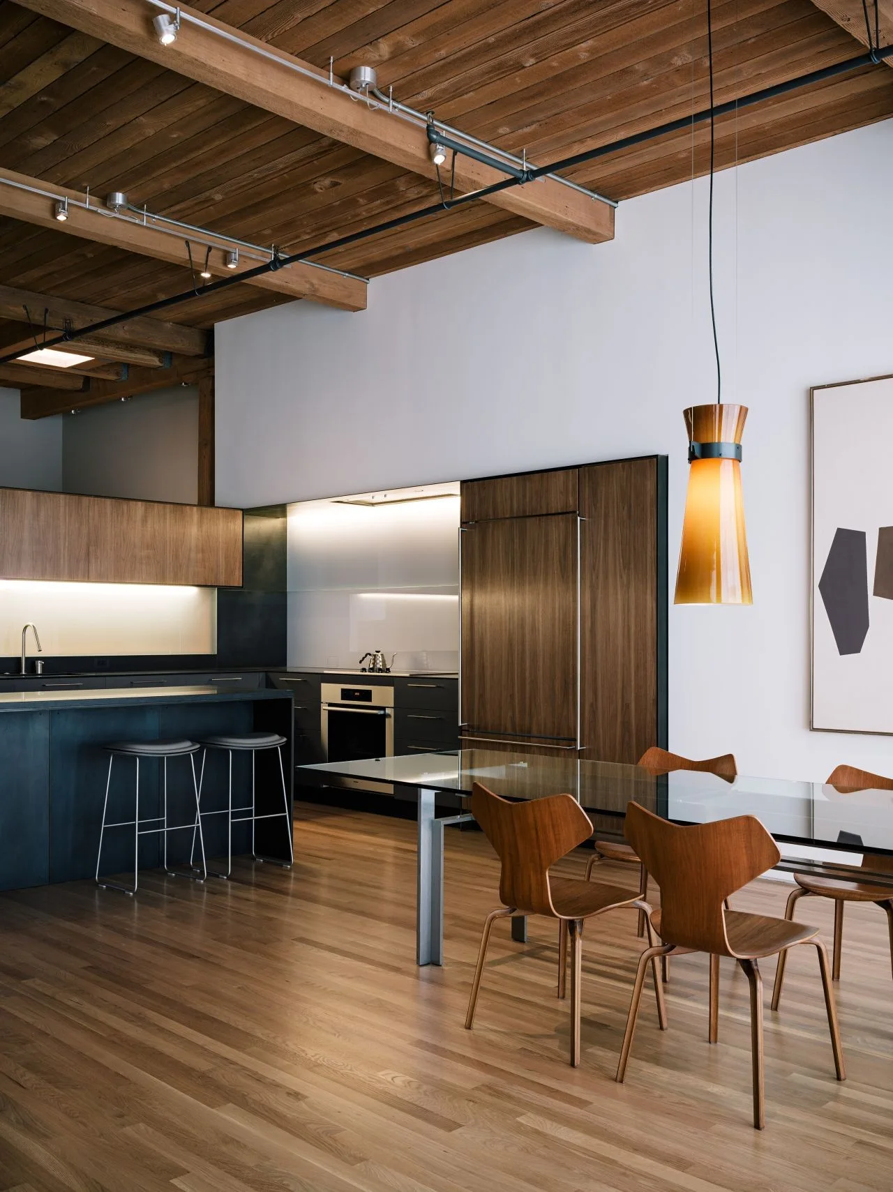 A modern open-plan kitchen and dining area with a glass table, mid-century wooden chairs, and dark cabinetry under an exposed wood ceiling.