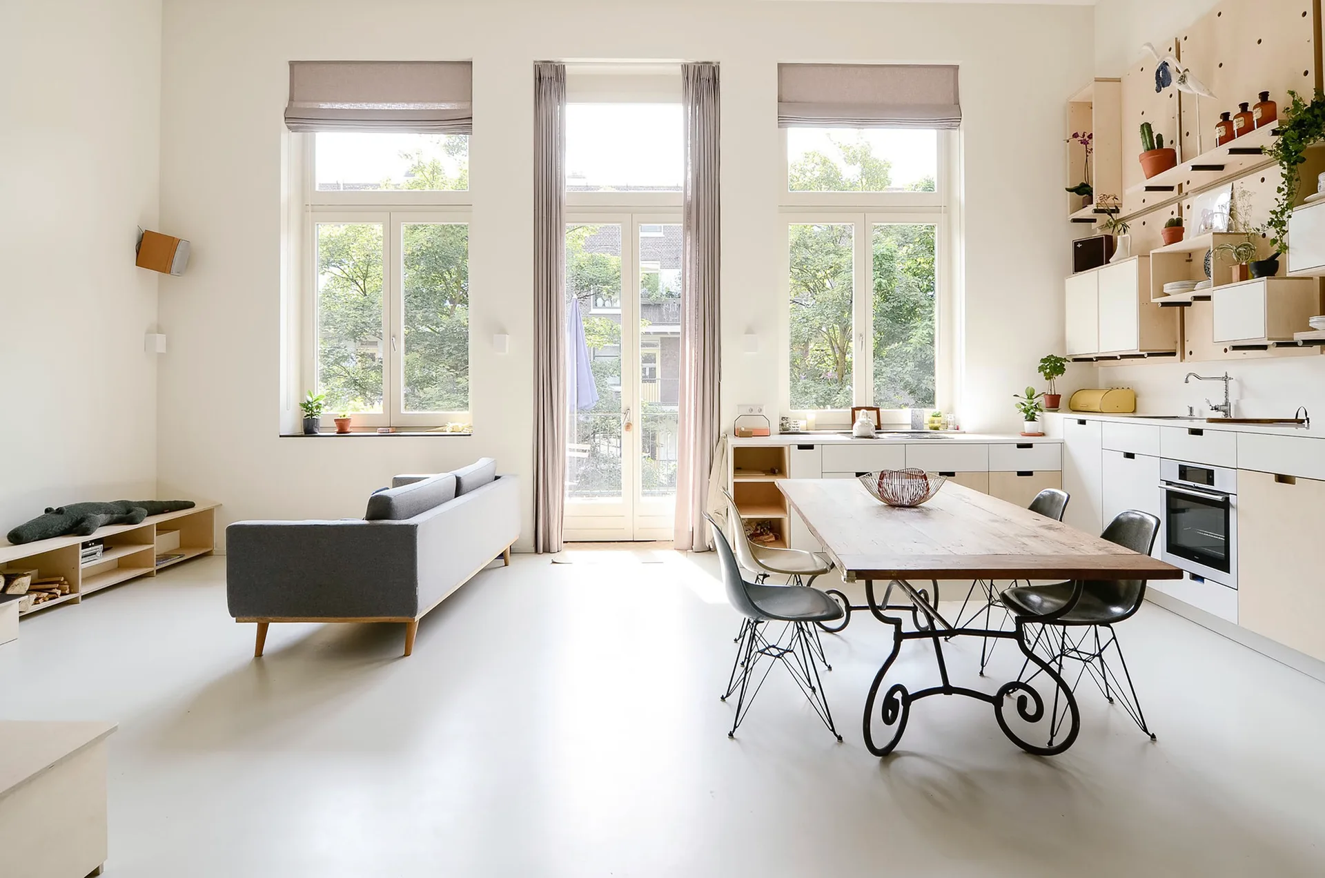 An open concept kitchen and living area with tall windows, a grey sofa, a wooden dining table, and a plywood pegboard wall system.