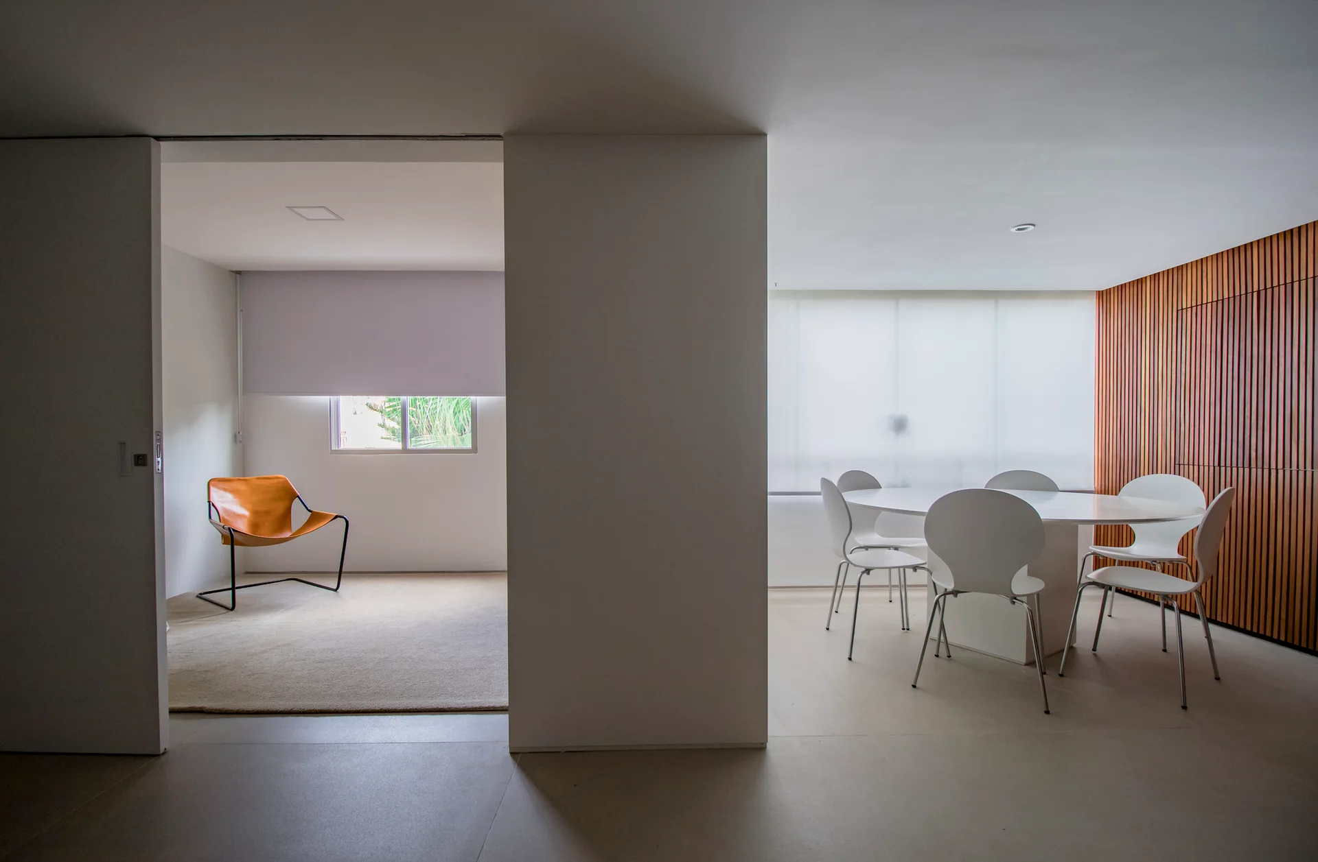 An open-concept modern interior displays a white dining table set beside a wood slat wall and an adjacent room with a leather sling chair.