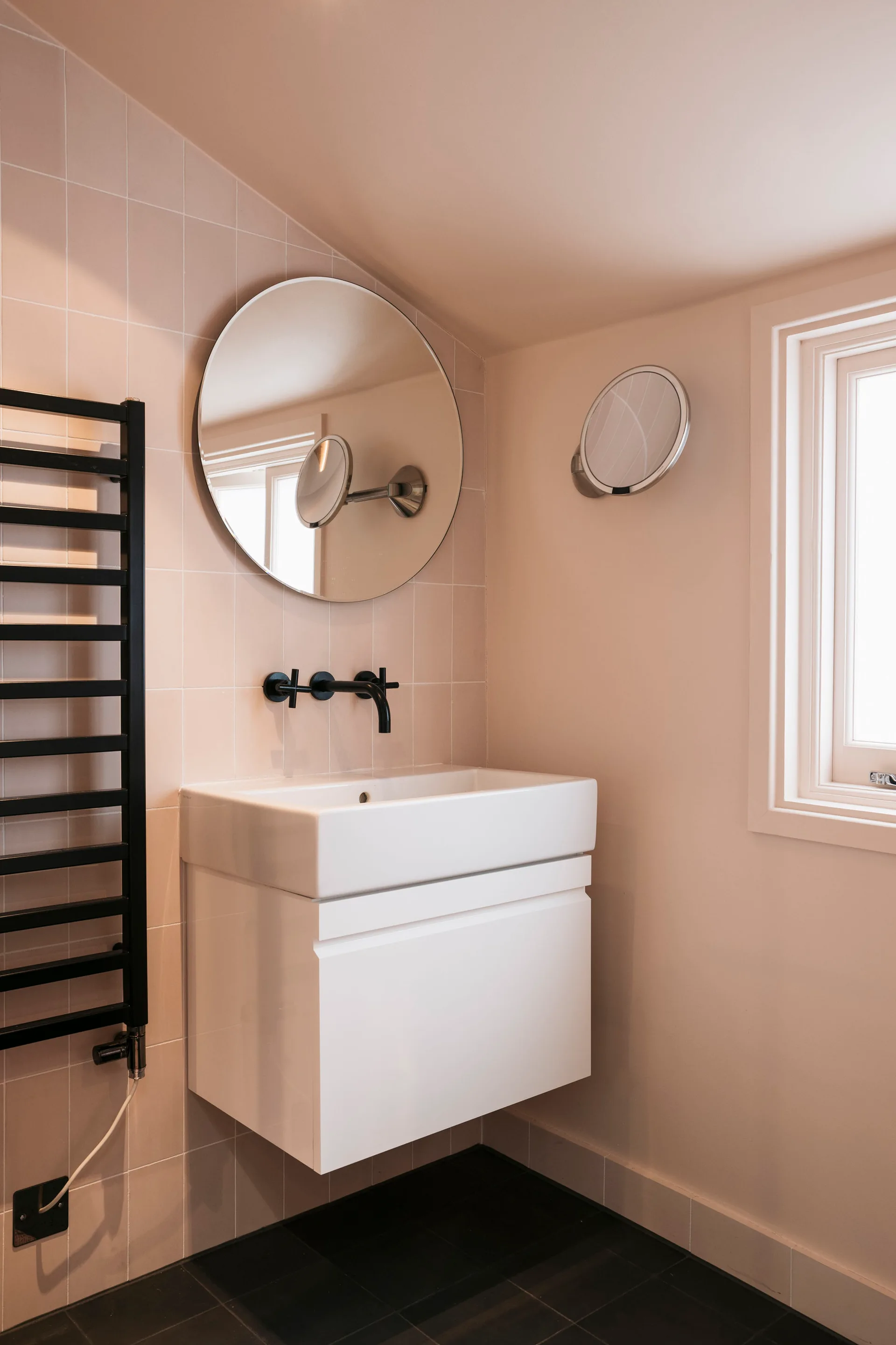 A minimalist bathroom featuring a wall-mounted white vanity sink, a circular mirror, and matte black fixtures against pale pink square wall tiles.