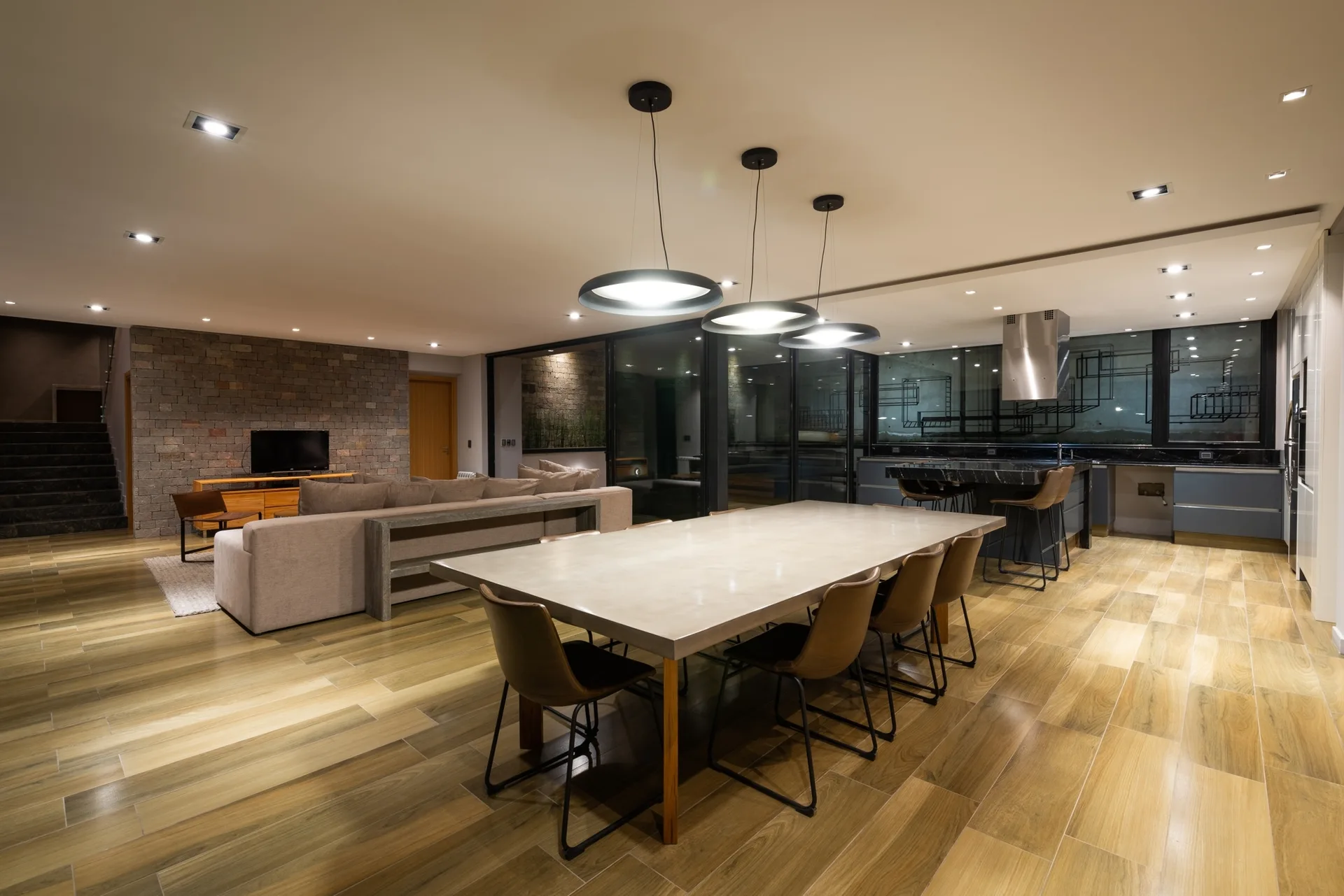 An open dining area features a concrete table with leather chairs under three disk pendant lights, with a living room and stone accent wall in the background.