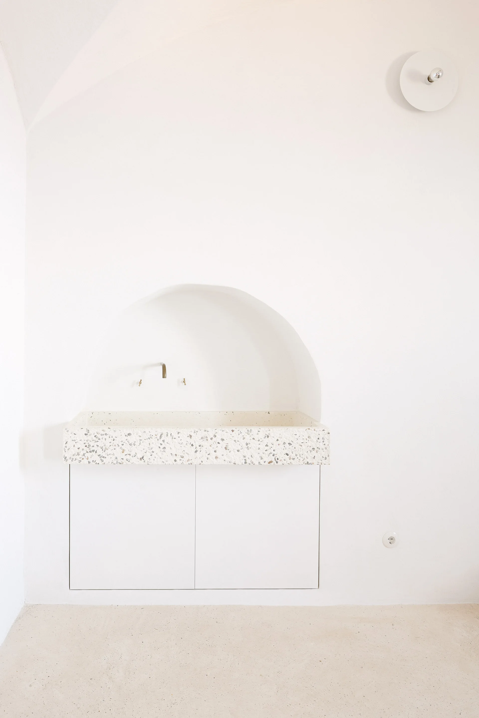A minimalist white bathroom featuring a built-in terrazzo vanity set beneath a recessed arched wall niche with a metal faucet.