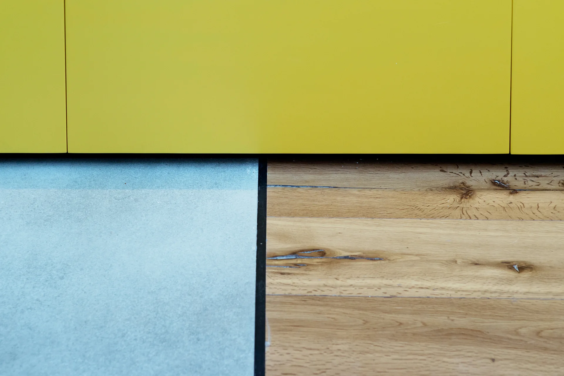 A close-up view showing the sharp junction where a bright yellow cabinet meets a grey textured rug and natural wood plank flooring.