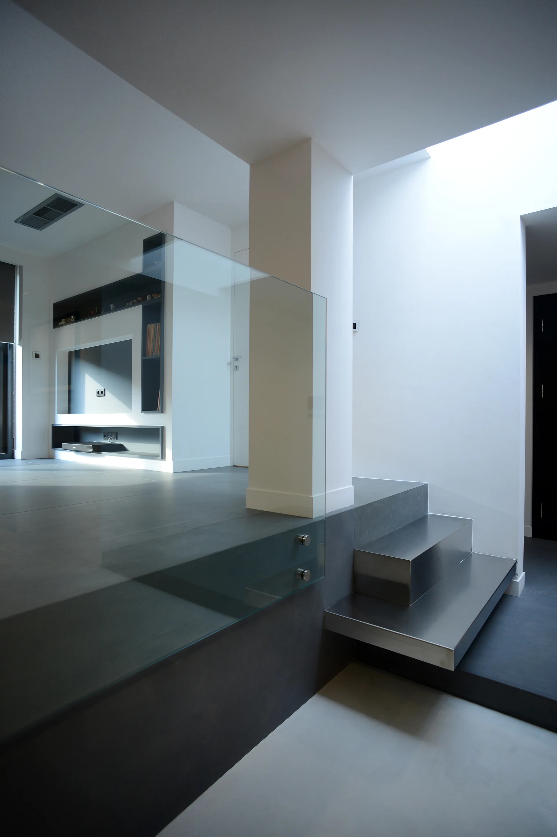 A minimalist split-level interior features a single metal step, a frameless glass railing, and a skylight casting light onto white walls.