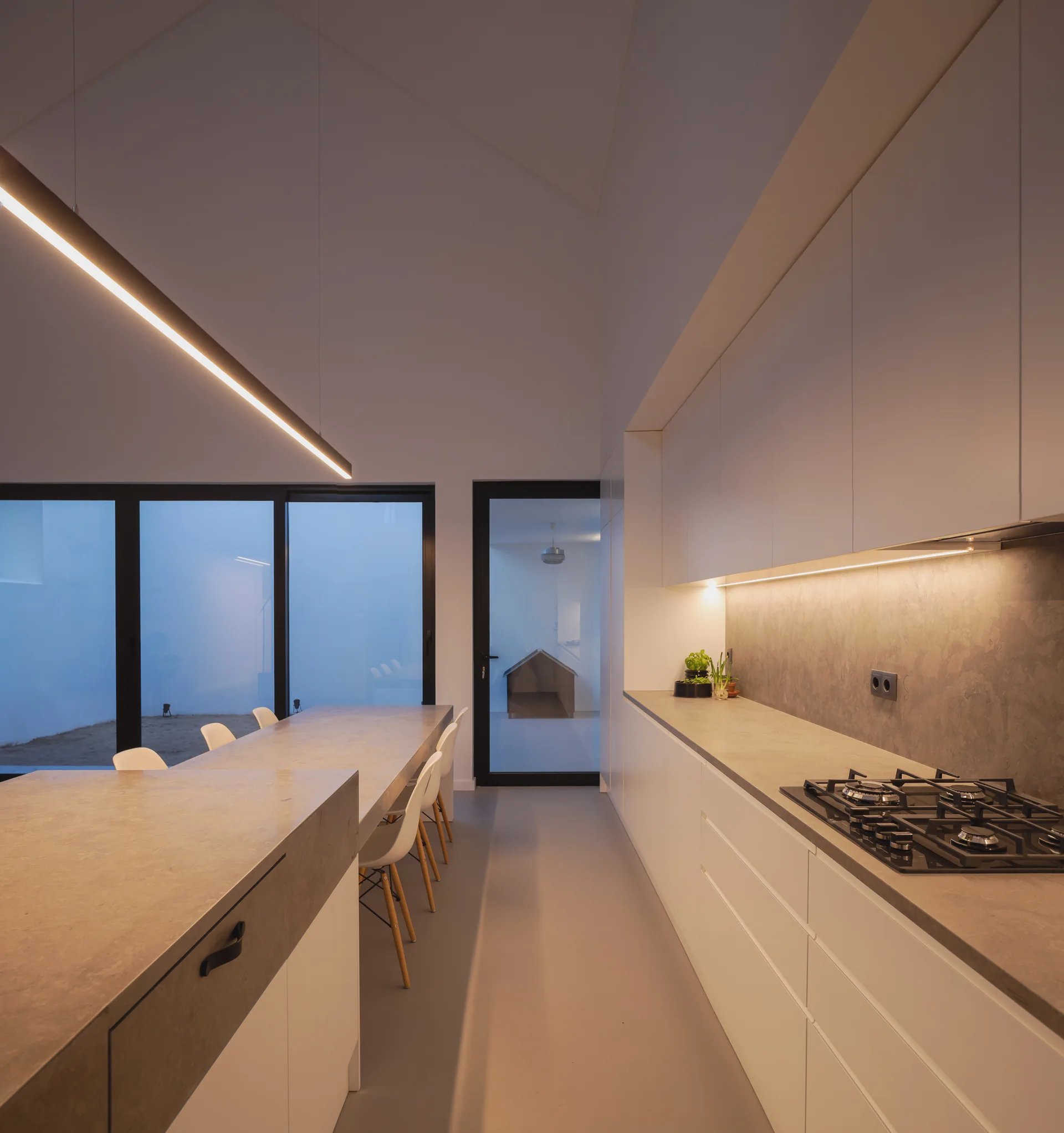A minimalist kitchen features flat-front cabinetry, stone-like countertops with a gas stovetop, and a long dining table under a vaulted ceiling.