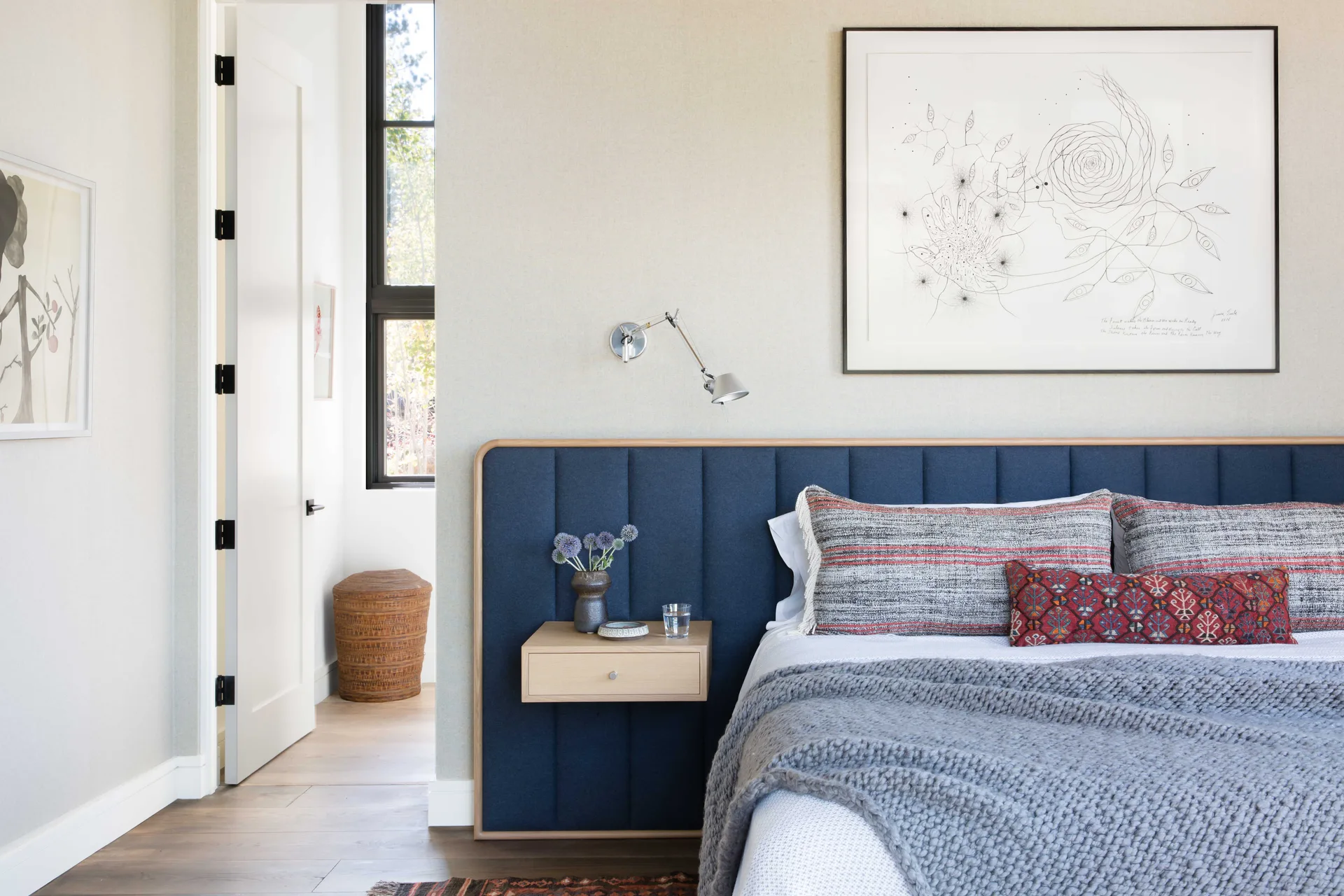 A modern bedroom features a deep blue upholstered headboard, a floating light wood nightstand, and a chunky grey knit throw blanket on the bed.