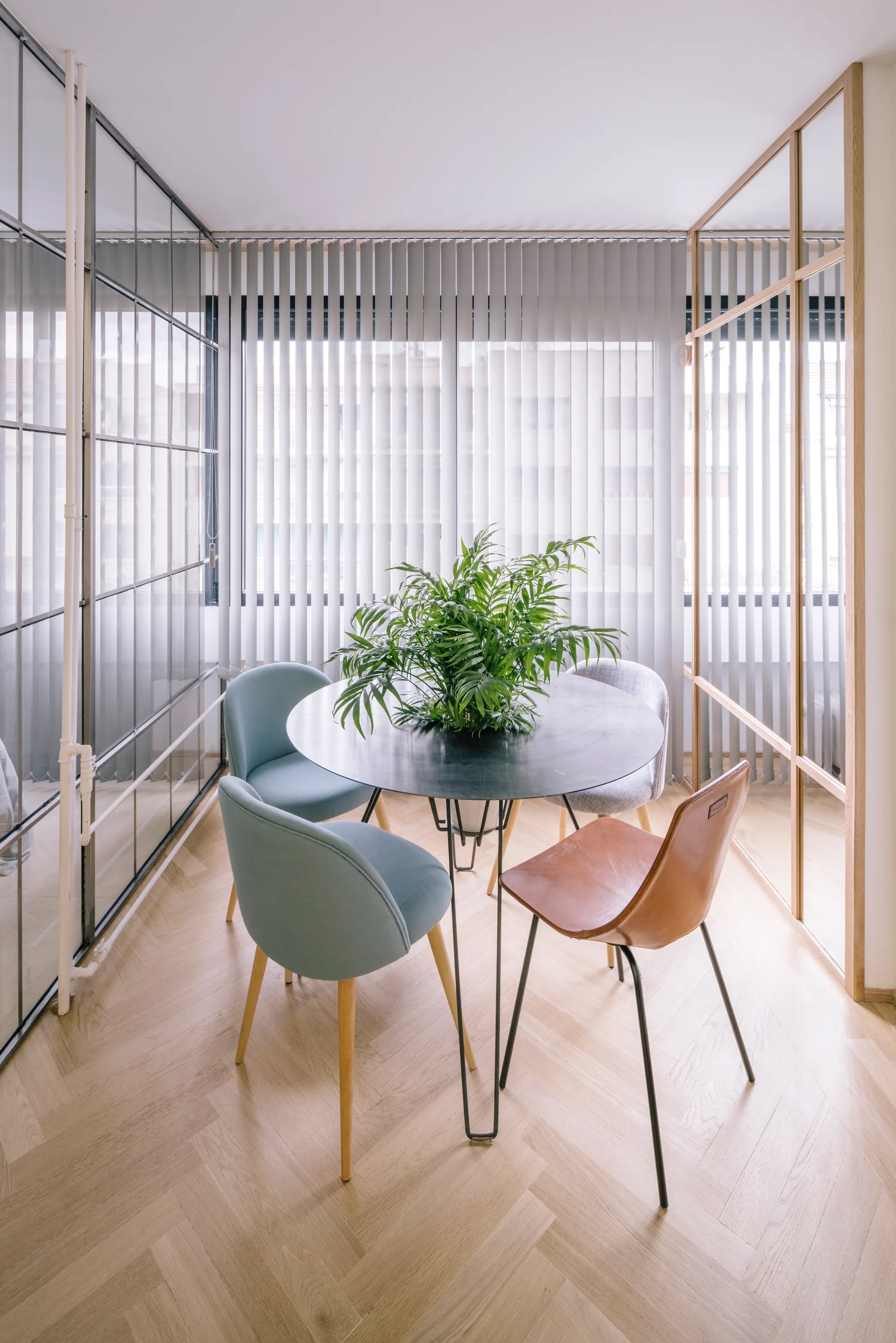 A round black dining table surrounded by mismatched chairs sits on herringbone wood flooring between glass partition walls.