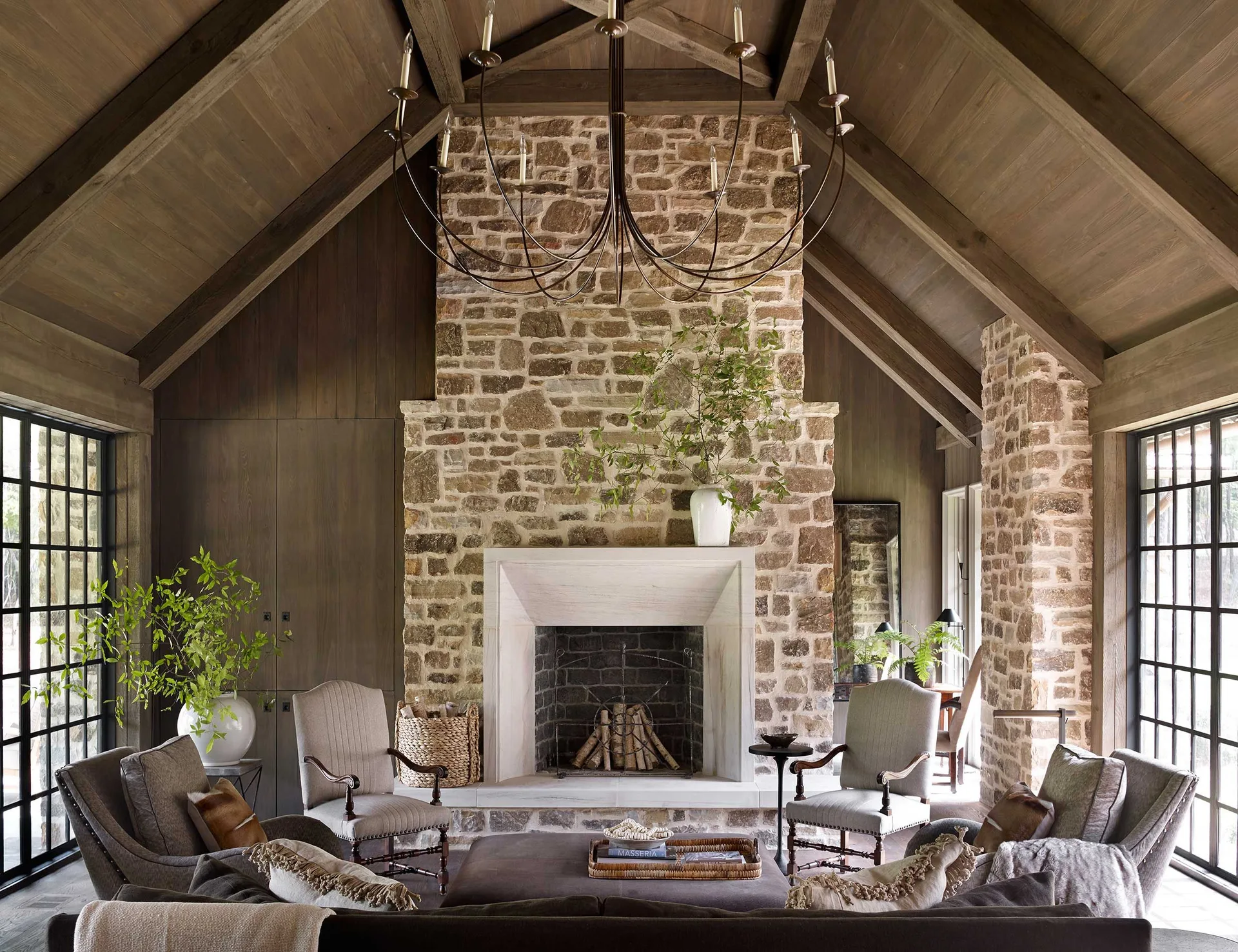 A rustic living room features a tall stone fireplace, a vaulted timber ceiling with exposed beams, and upholstered armchairs.