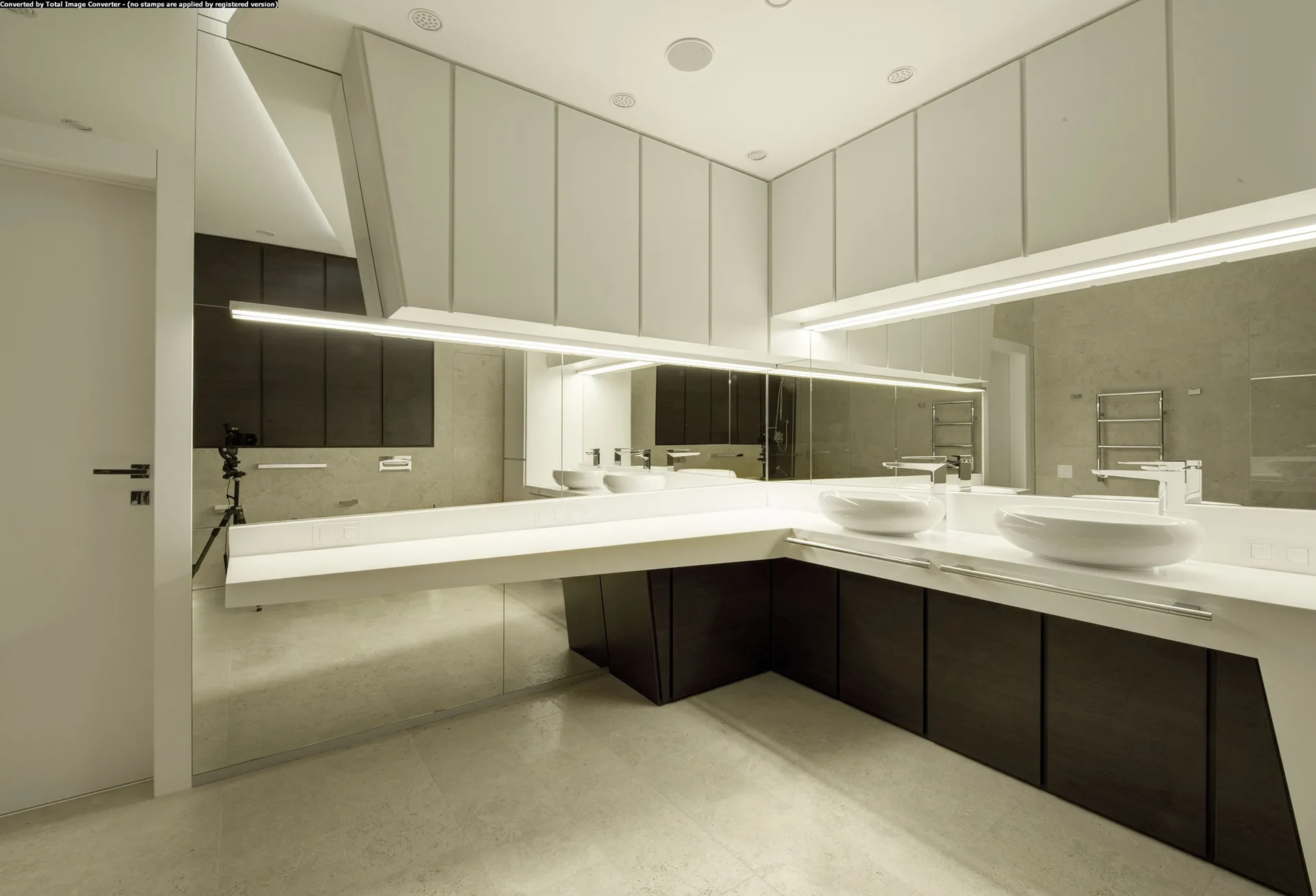 A minimalist bathroom featuring a dark wood L-shaped floating vanity with two white vessel sinks and a large wall-spanning mirror.