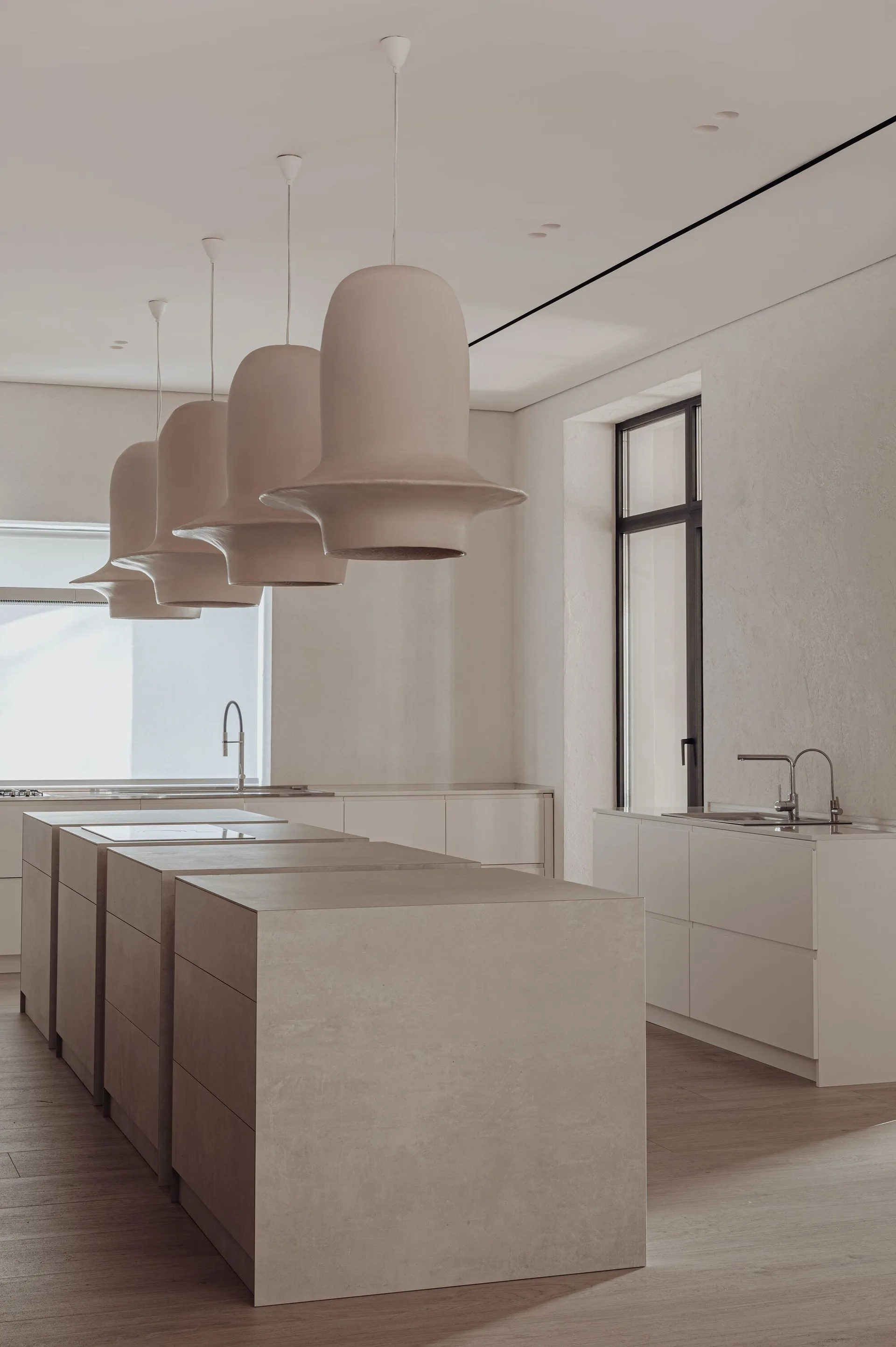 A minimalist kitchen featuring four large sculptural beige pendant lights hanging over monolithic block islands.