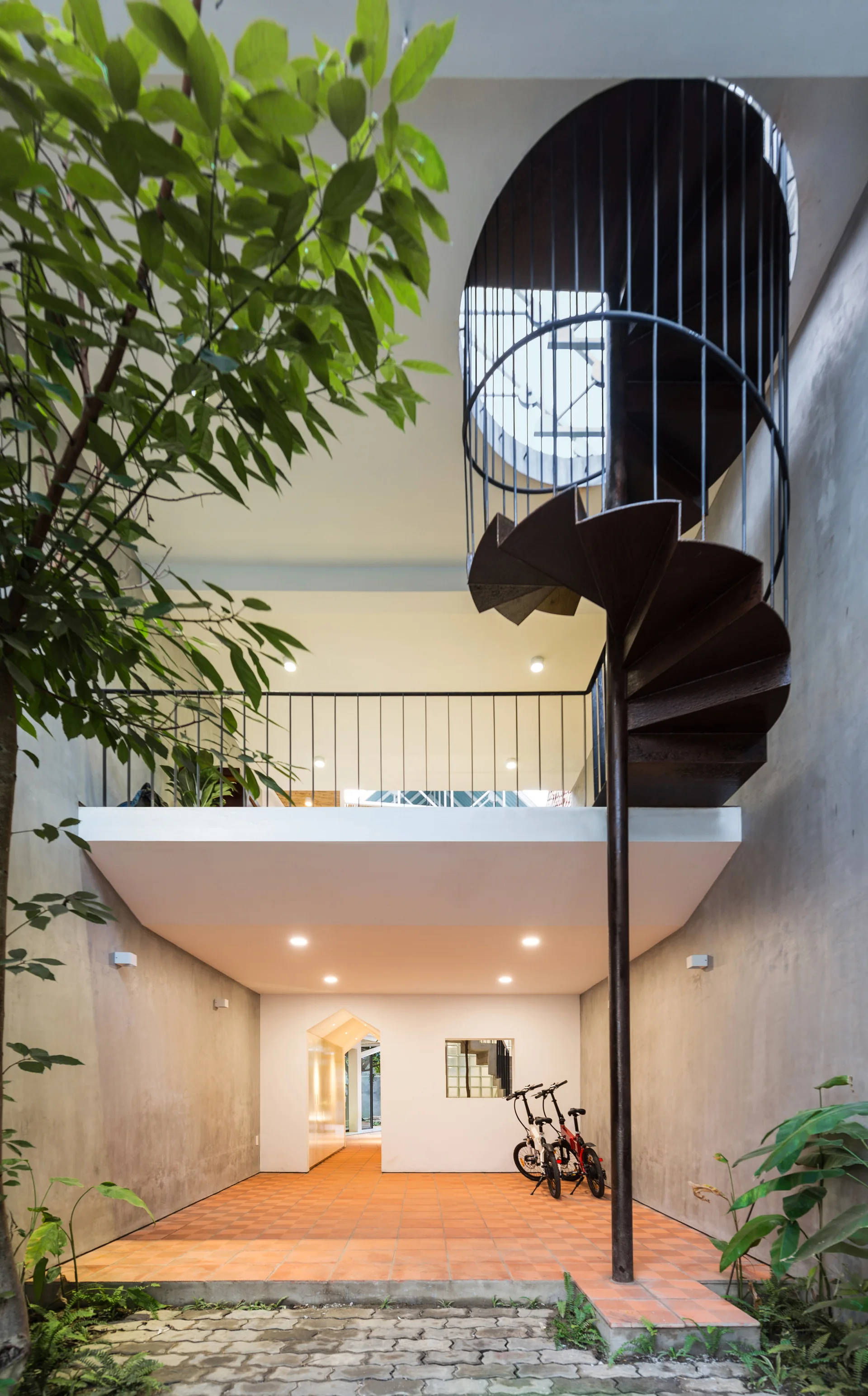 A black metal spiral staircase rises from a terracotta tiled floor within an open concrete courtyard atrium with a mezzanine level.