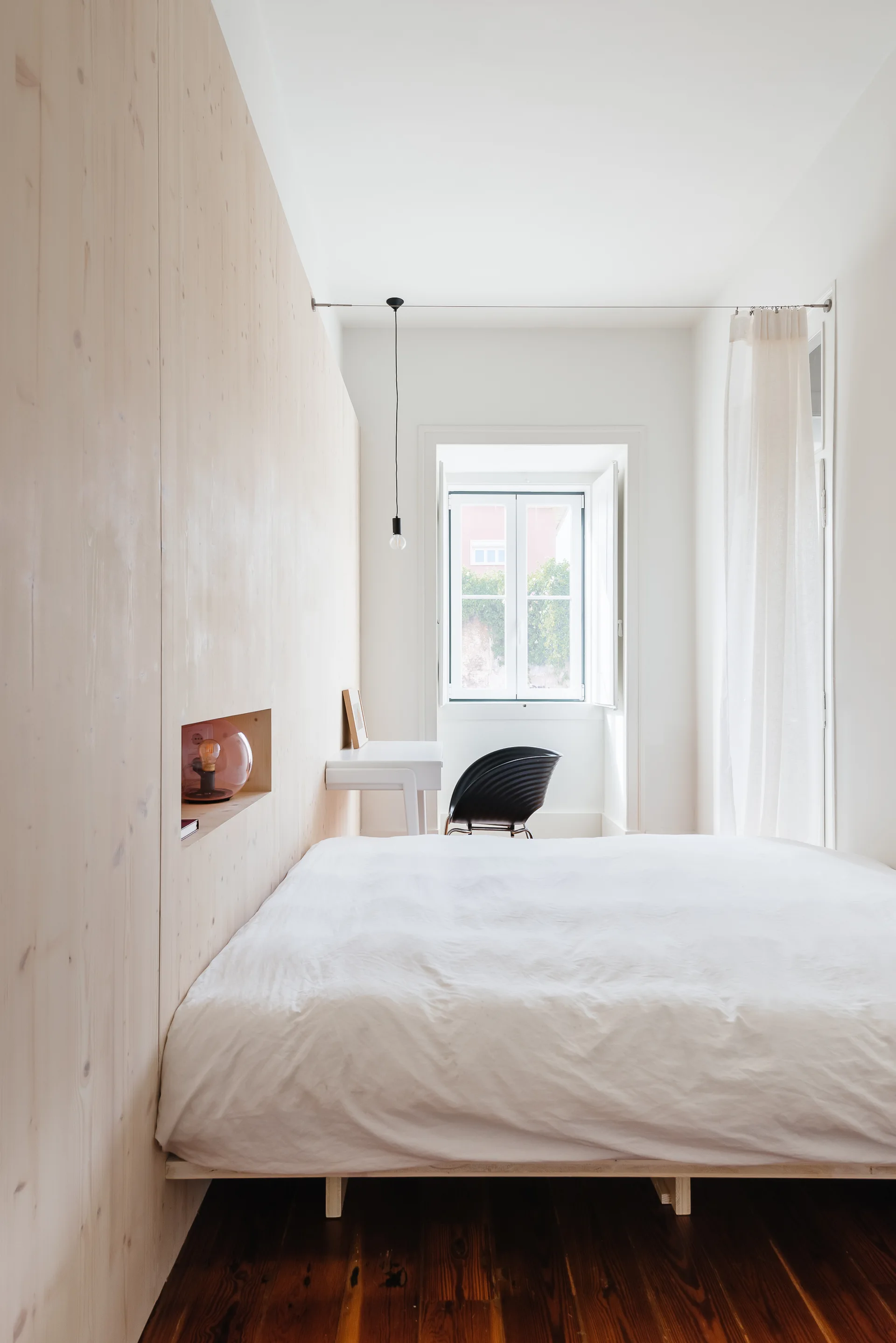 A minimalist bedroom featuring a bed beside a light wood paneled wall with a small built-in niche, a white desk, and a window.