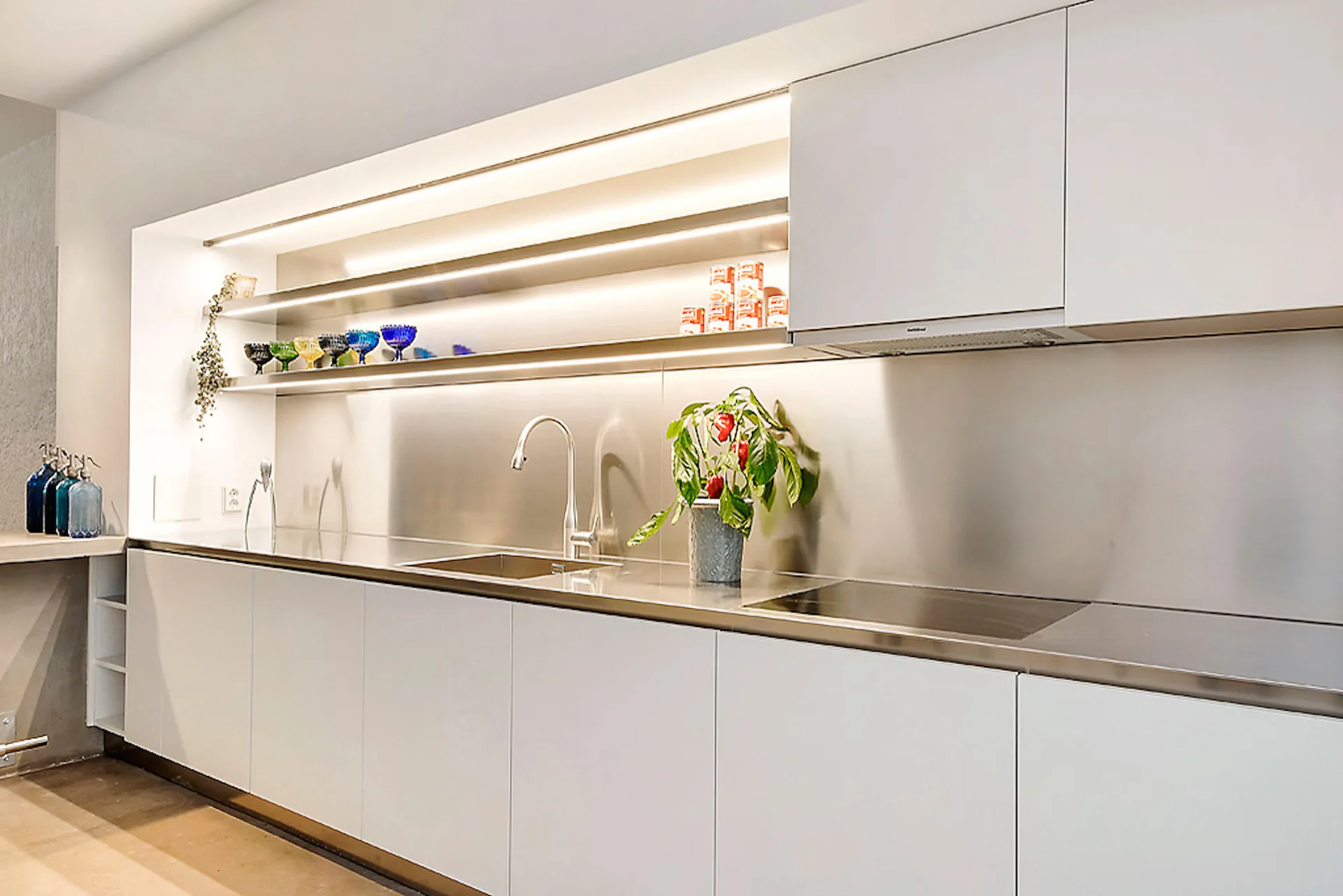 A modern kitchen featuring white handleless cabinetry beneath a continuous stainless steel countertop and backsplash with integrated lighted shelving.