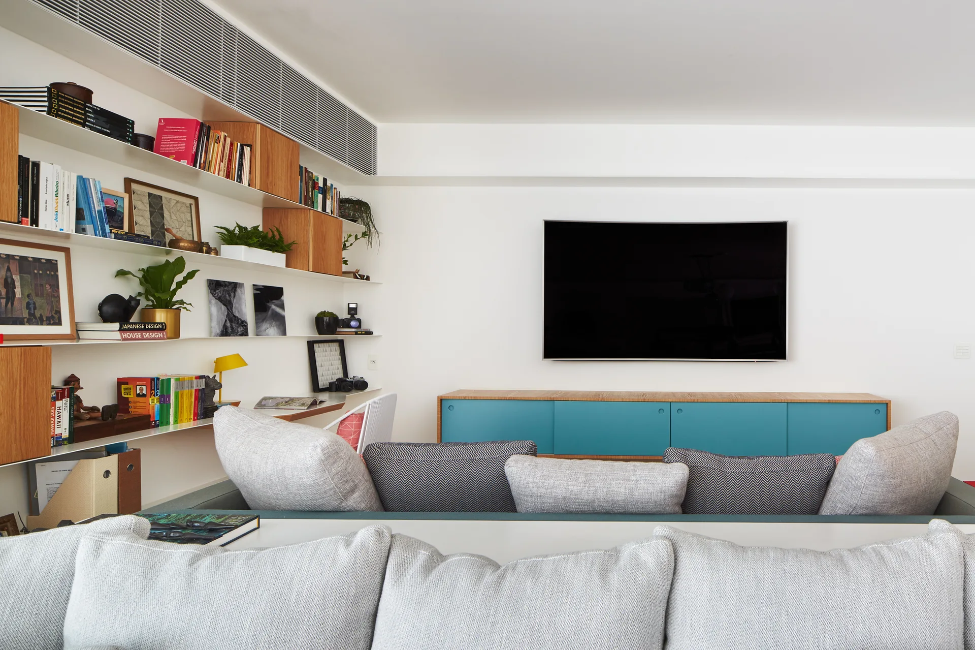 A grey sofa sits in front of a wall-mounted TV, a blue storage console, and white floating wall shelves filled with books and plants.