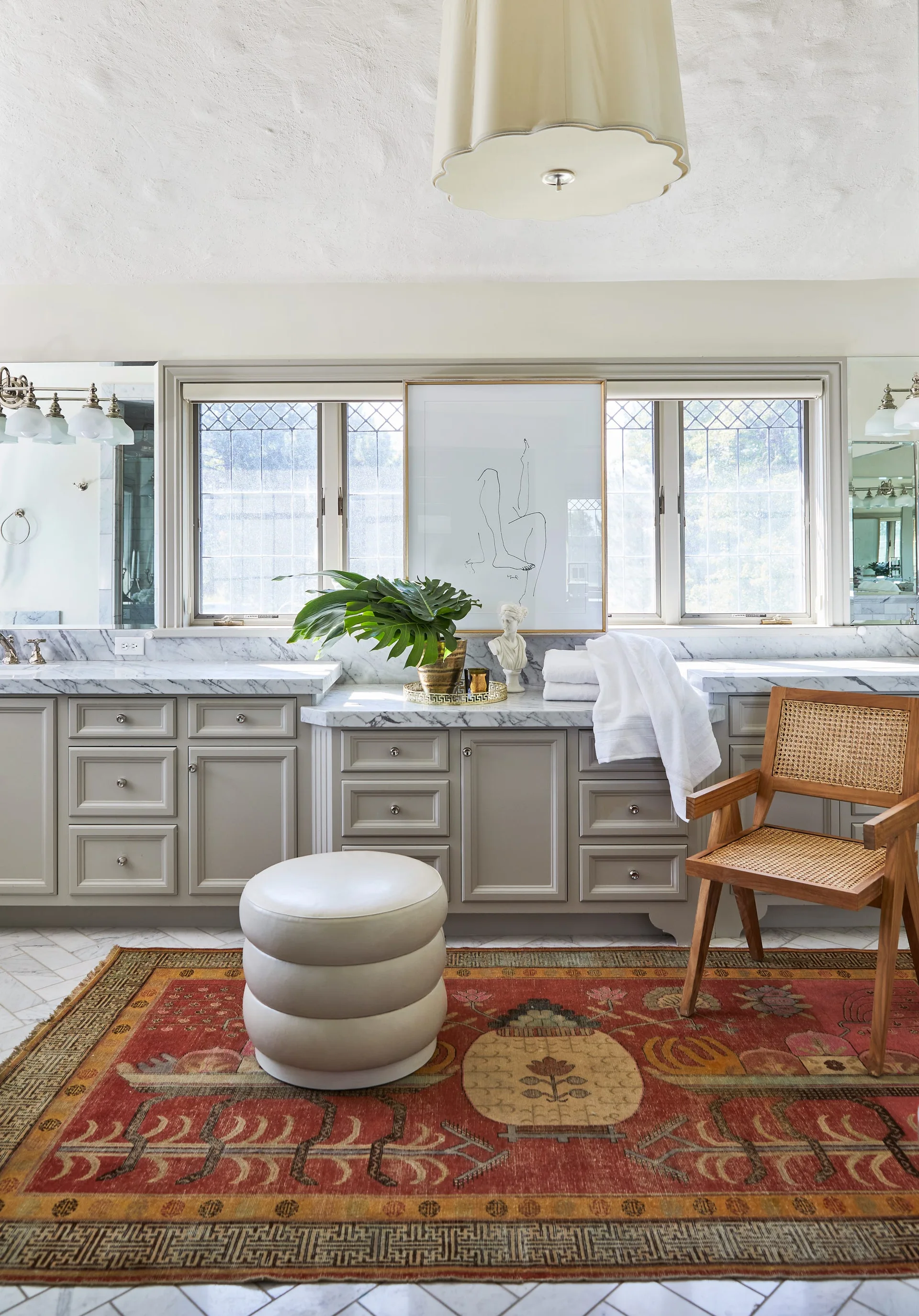 A grey bathroom vanity with a marble countertop features leaded windows, a stacked round ottoman, and a wooden cane chair on a red patterned rug.