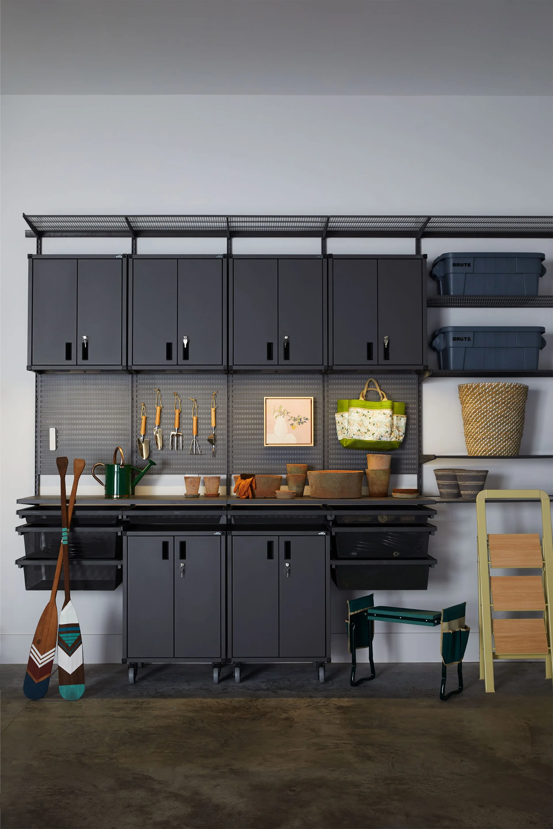 A wall-mounted matte black metal storage system features upper cabinets, a pegboard with tools, lower rolling cabinets, and a yellow step ladder.