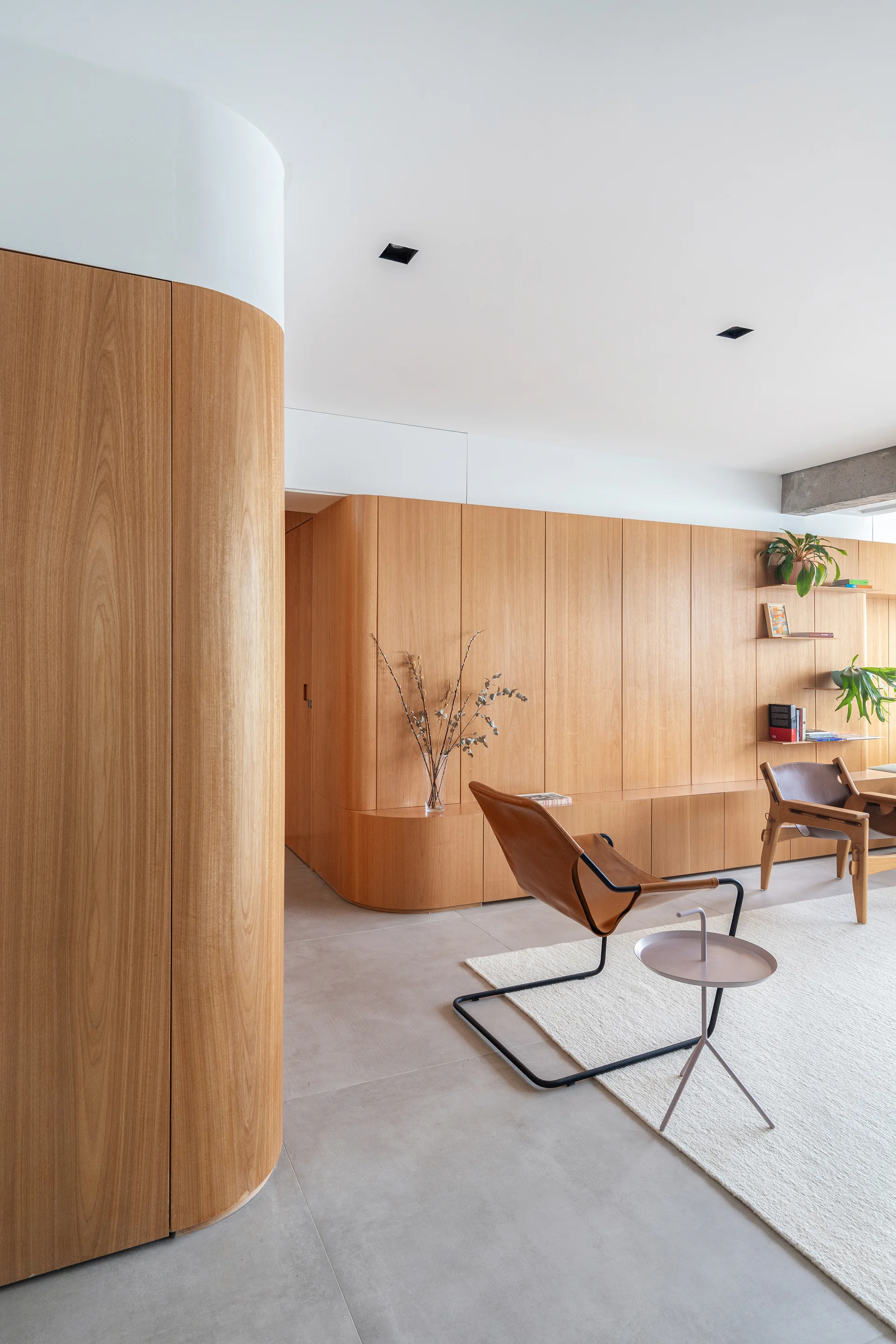 A modern living area features curved light wood cabinetry, tan leather lounge chairs, and grey concrete flooring under a smooth white ceiling.