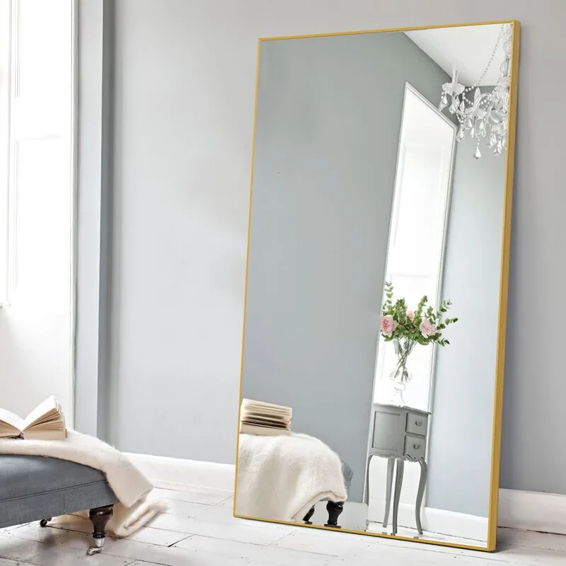 A large rectangular floor mirror with a thin gold frame leans against a light grey wall next to a grey bench with a white throw blanket.