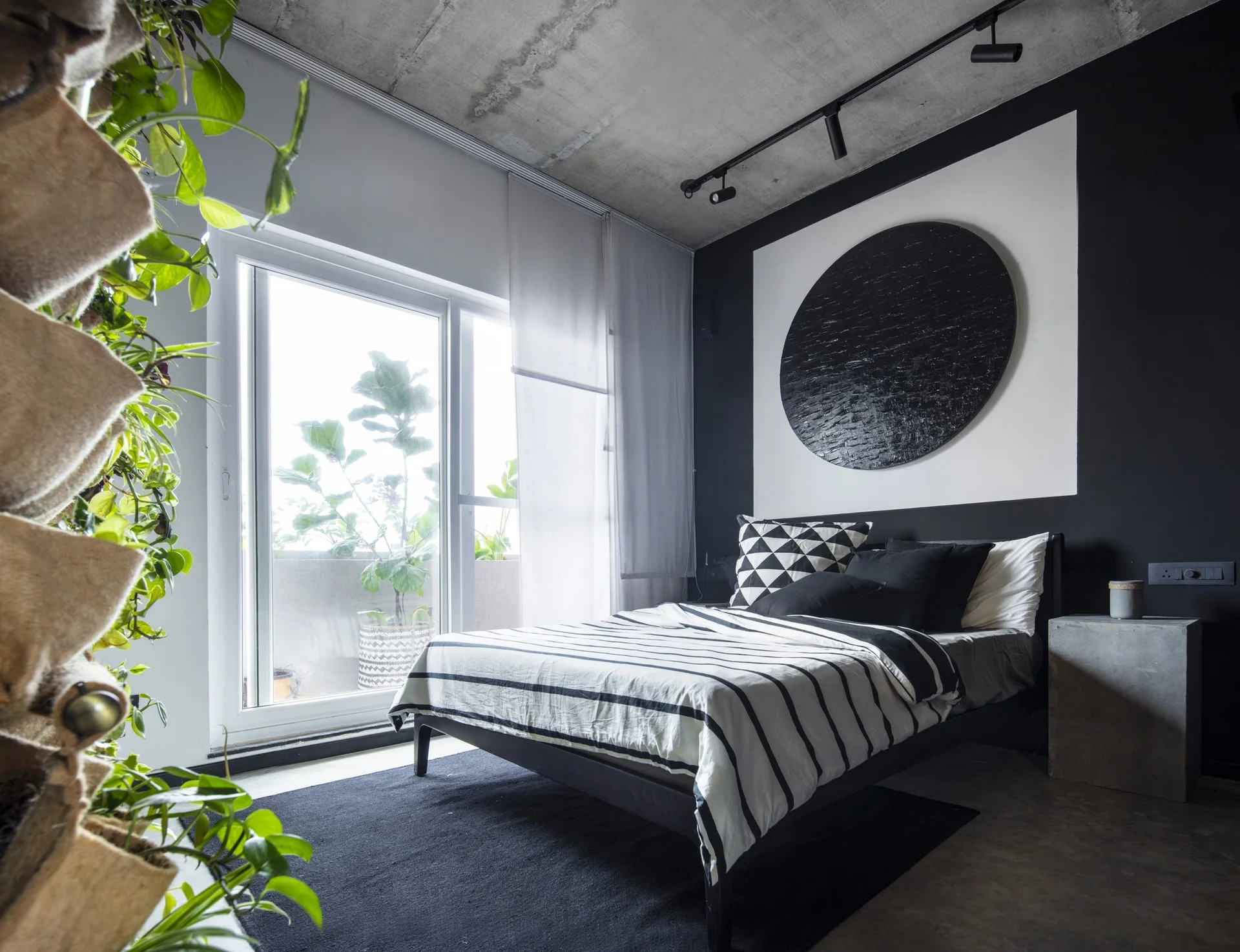 A modern bedroom features a bed with striped bedding under a raw concrete ceiling, a large black circular art piece, and sliding glass doors.