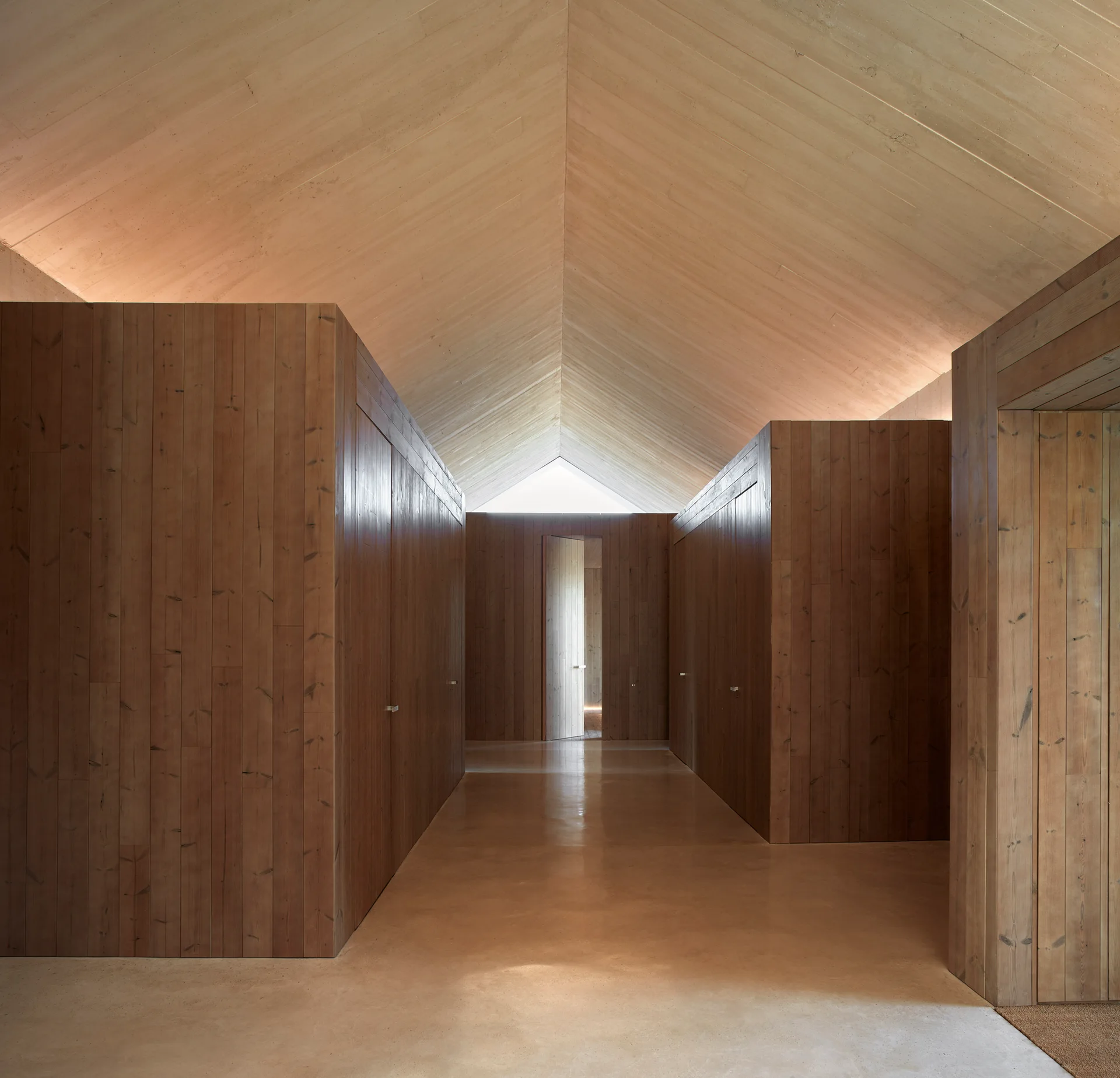A minimalist hallway features tall wood-paneled walls, a matching gabled wood ceiling, and a light beige polished concrete floor.