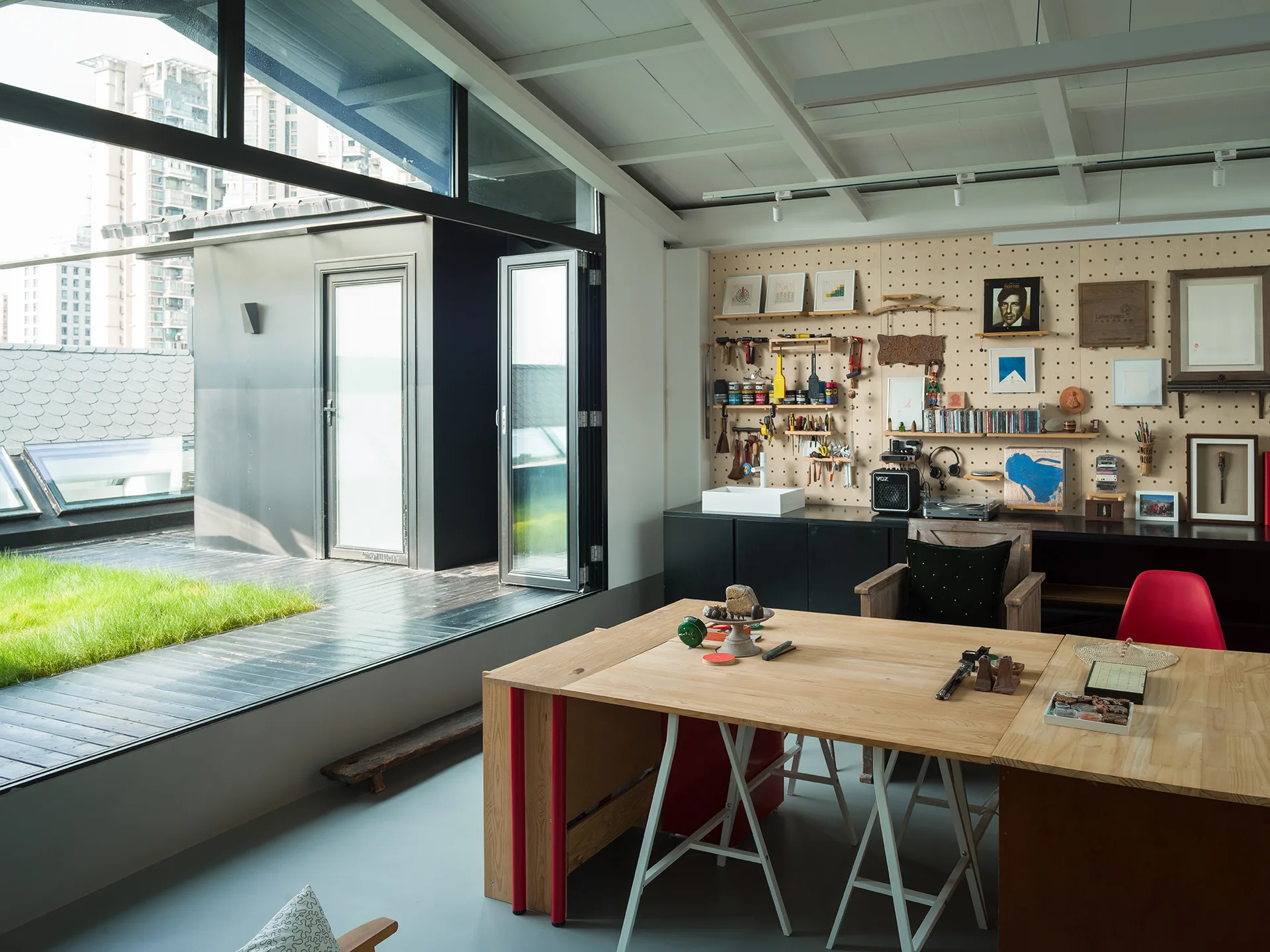 A light-filled creative workspace features a large wooden desk, a pegboard storage wall, and folding glass doors that open to a grassy outdoor terrace.