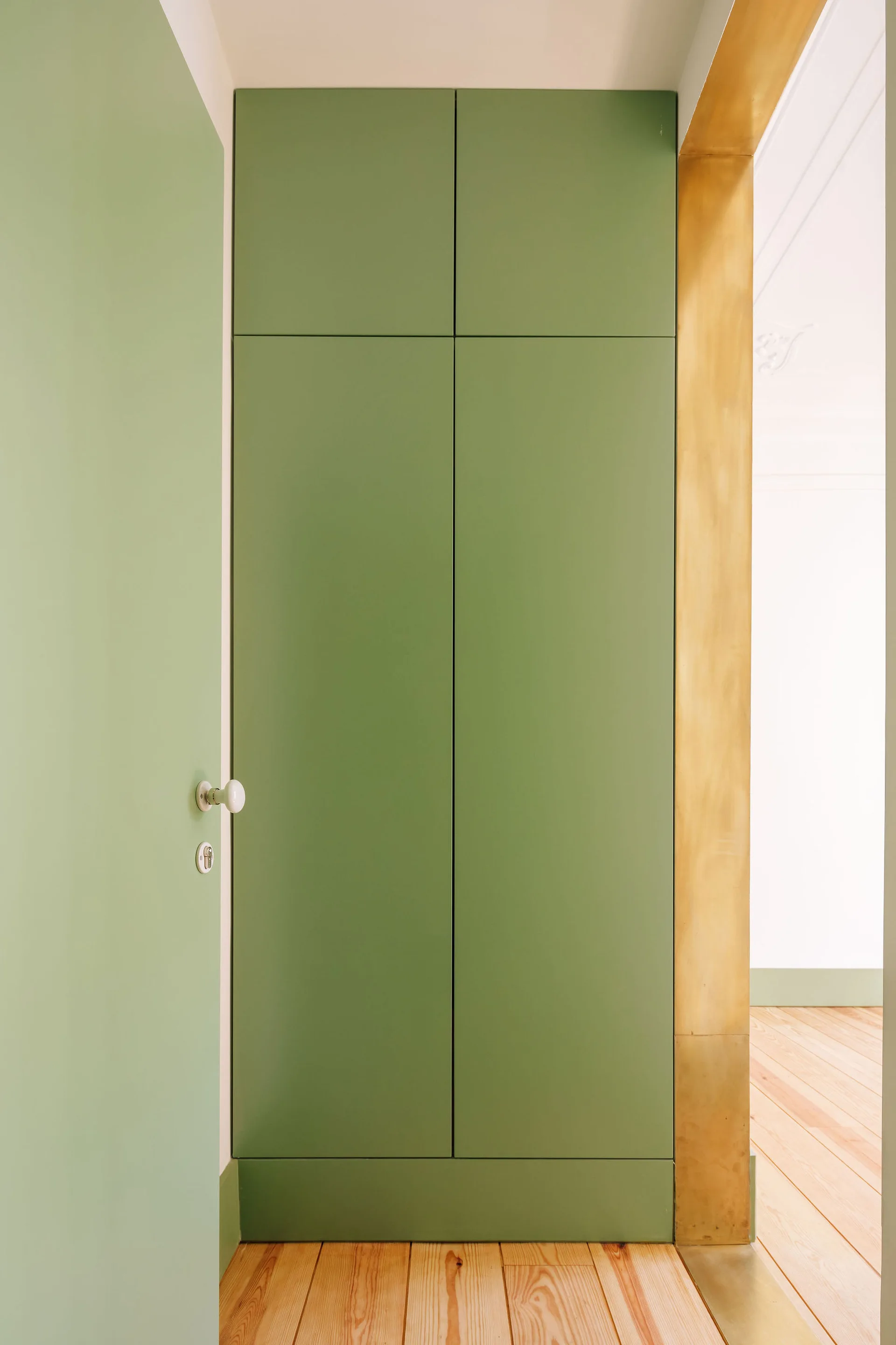 Full-height sage green cabinets stand next to a doorway framed in warm brass trim over light wood flooring.
