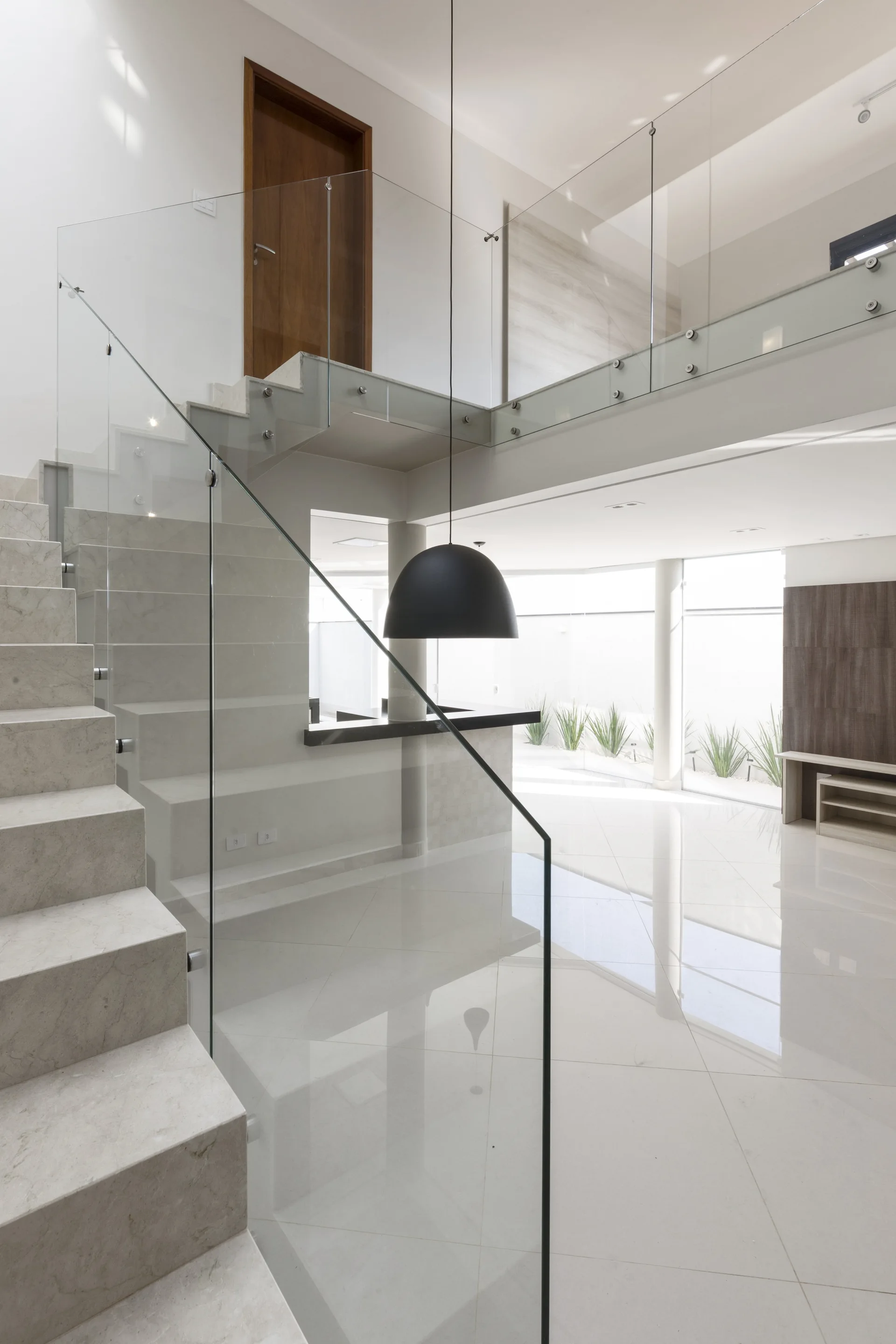 A light gray stone staircase with a frameless glass railing stands in a bright foyer with polished white tile floors and a black dome pendant light.