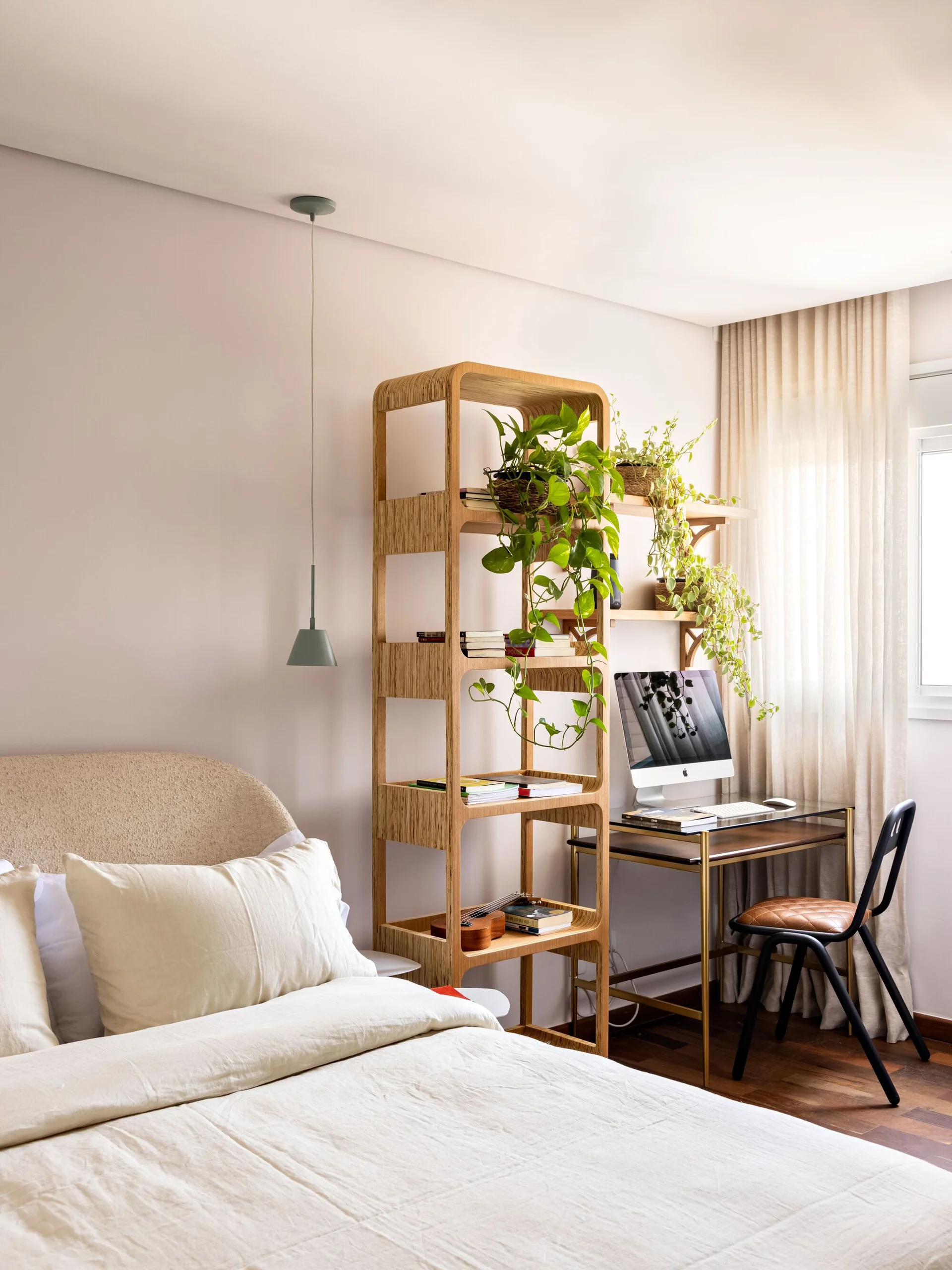 A bedroom features a bed next to a tall open plywood bookshelf and a small study desk in front of a window.