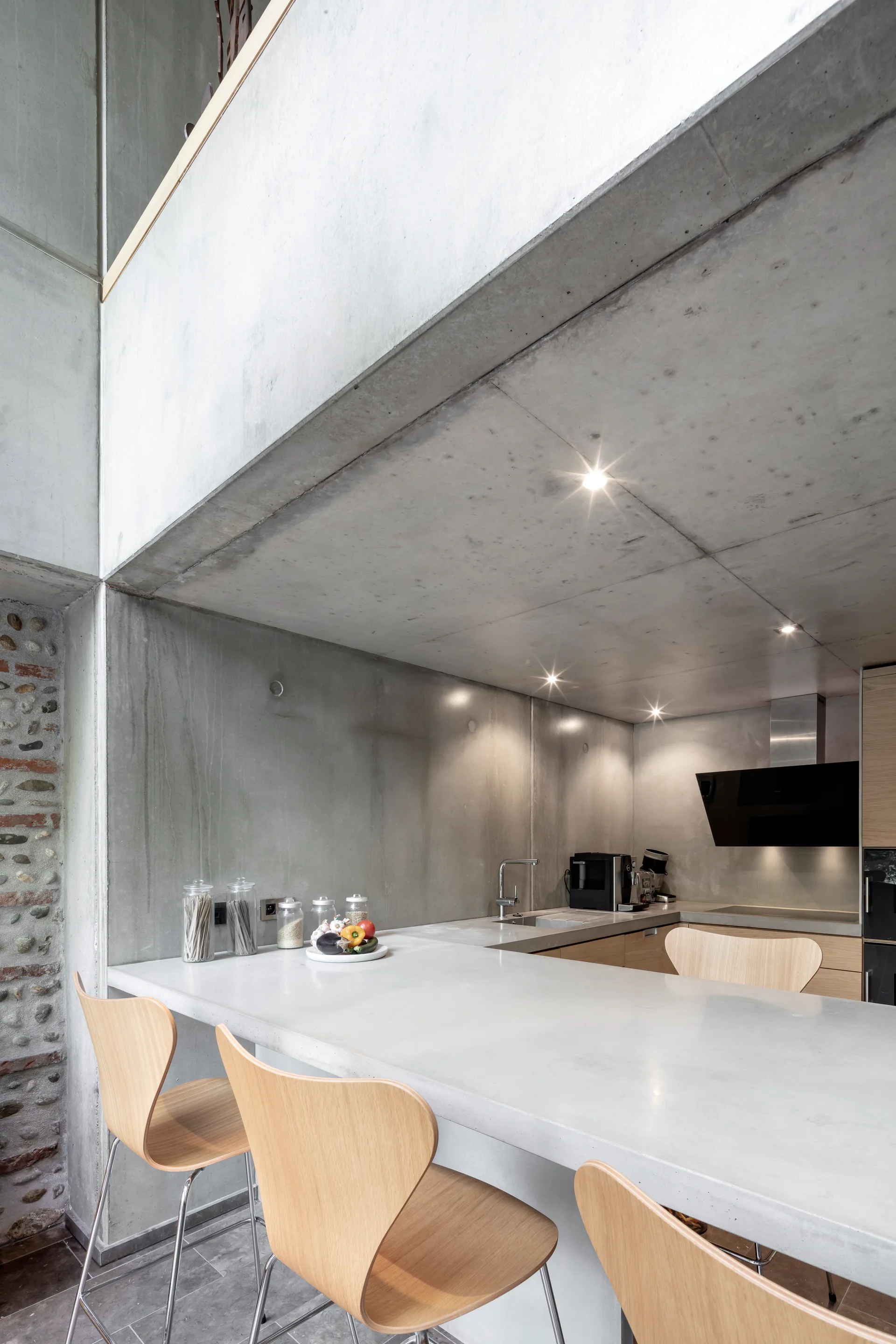A modern industrial kitchen features a smooth concrete breakfast bar paired with light wood bar stools under an exposed concrete ceiling.