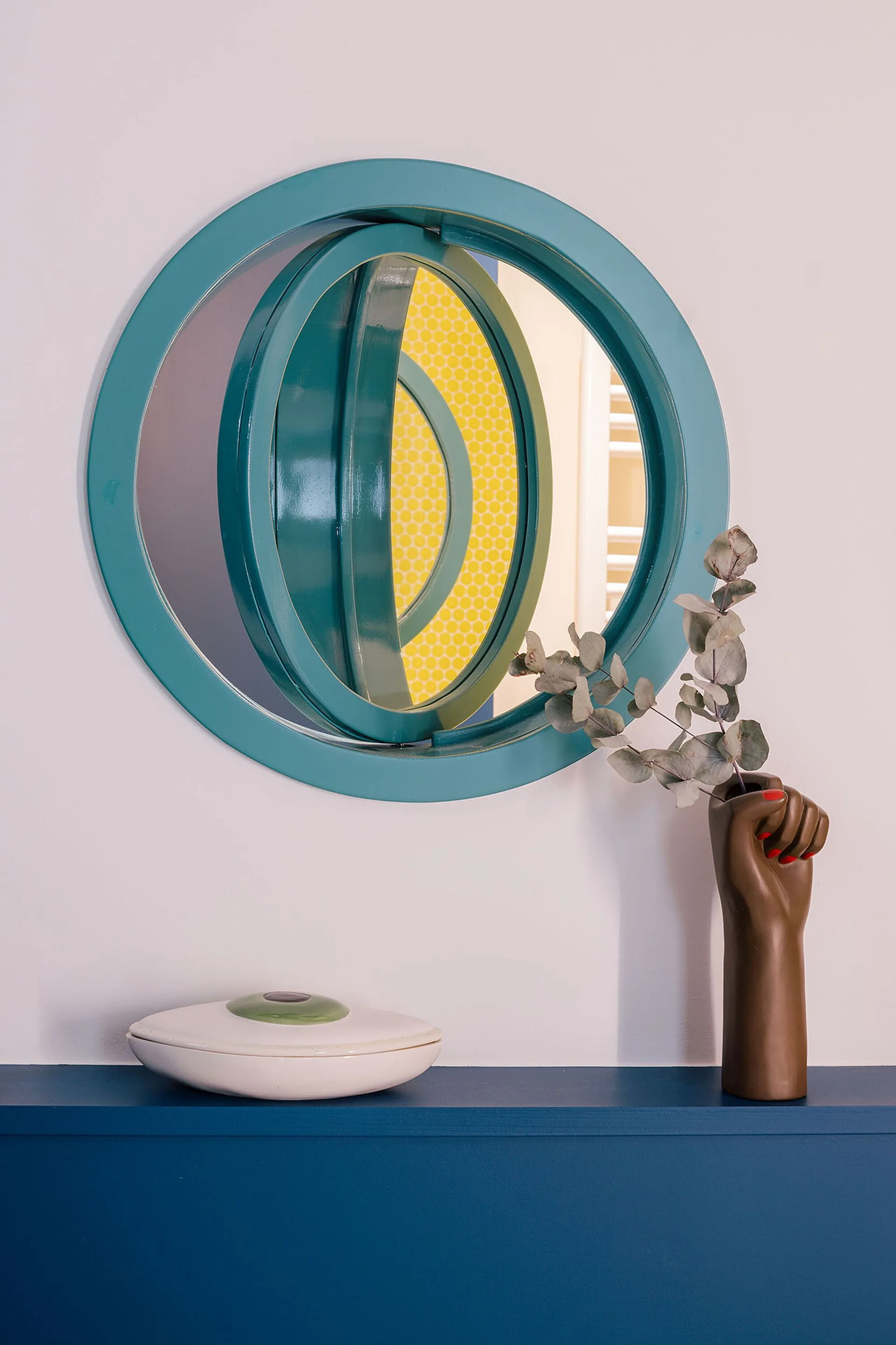 A circular teal mirror hangs on a white wall above a blue shelf displaying a brown fist-shaped ceramic vase with eucalyptus and a white lidded dish.