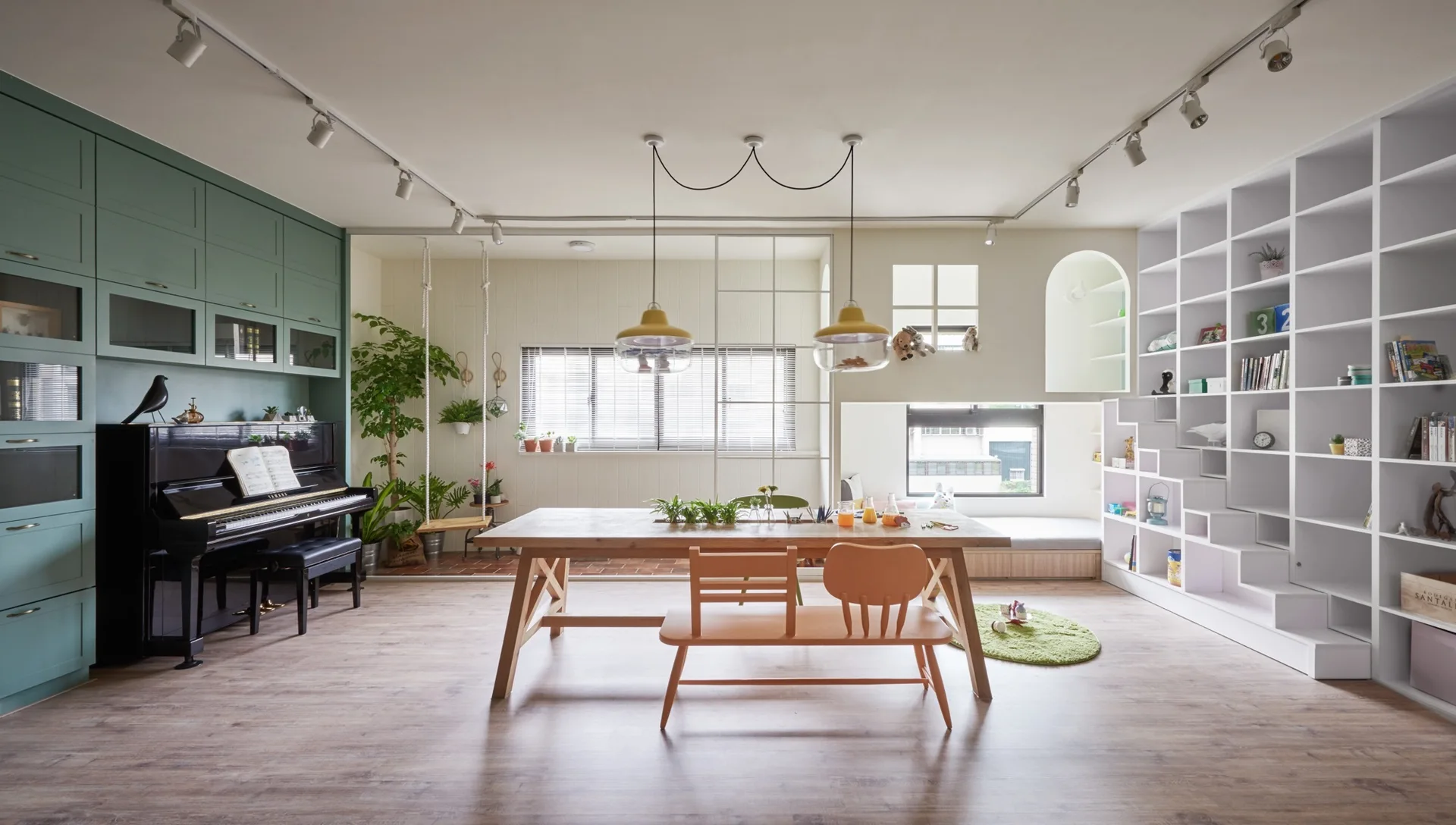 A bright family room features a wooden dining table with a peach bench, a black piano beneath sage green cabinets, and a massive white built-in bookshelf.