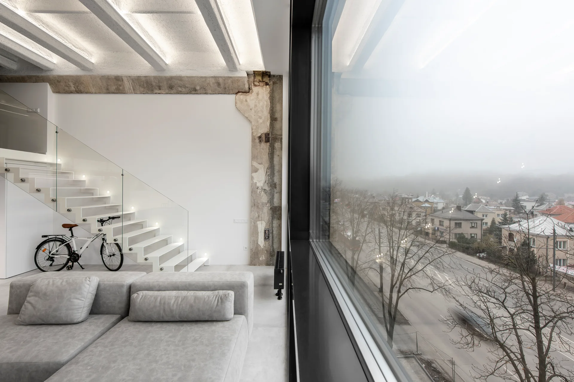 A minimalist grey sofa sits near a floating staircase with glass railings and an exposed concrete column next to a large window.