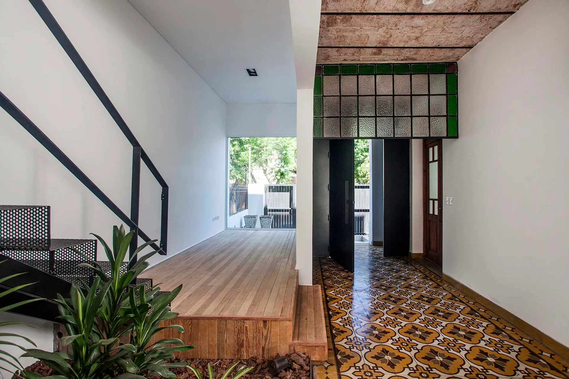 A modern entryway features patterned yellow tile flooring, a raised wood platform, white walls, and a black perforated metal staircase.