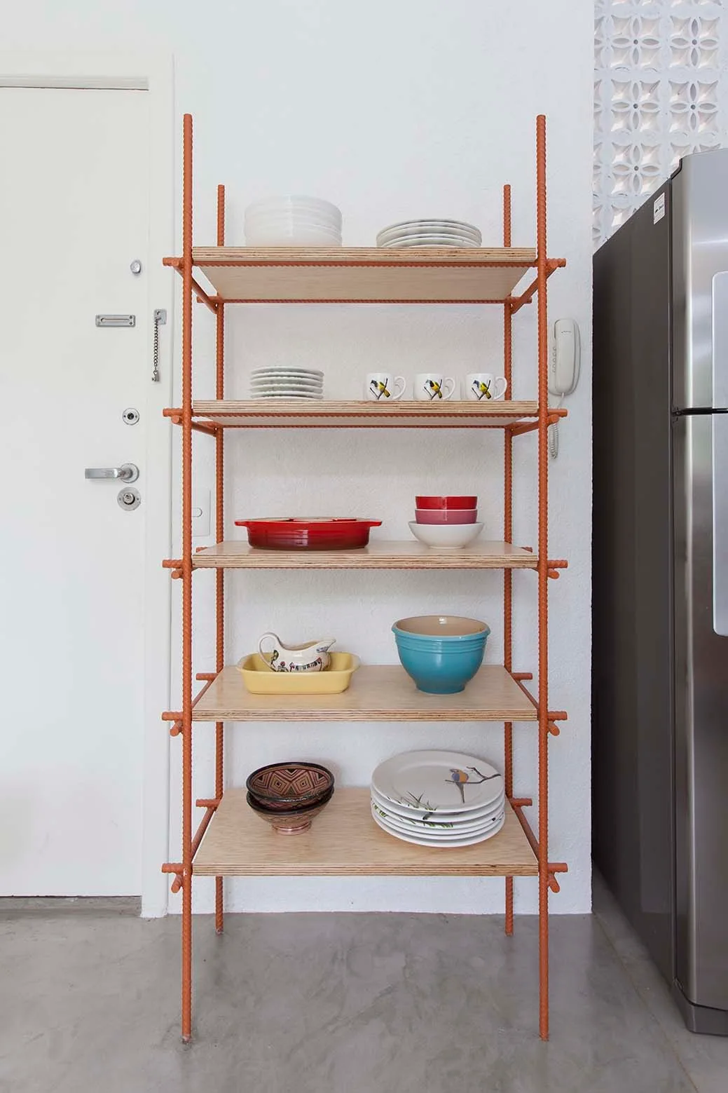An open shelving unit made of orange rebar and plywood holds colorful dishes against a white kitchen wall next to a stainless steel refrigerator.