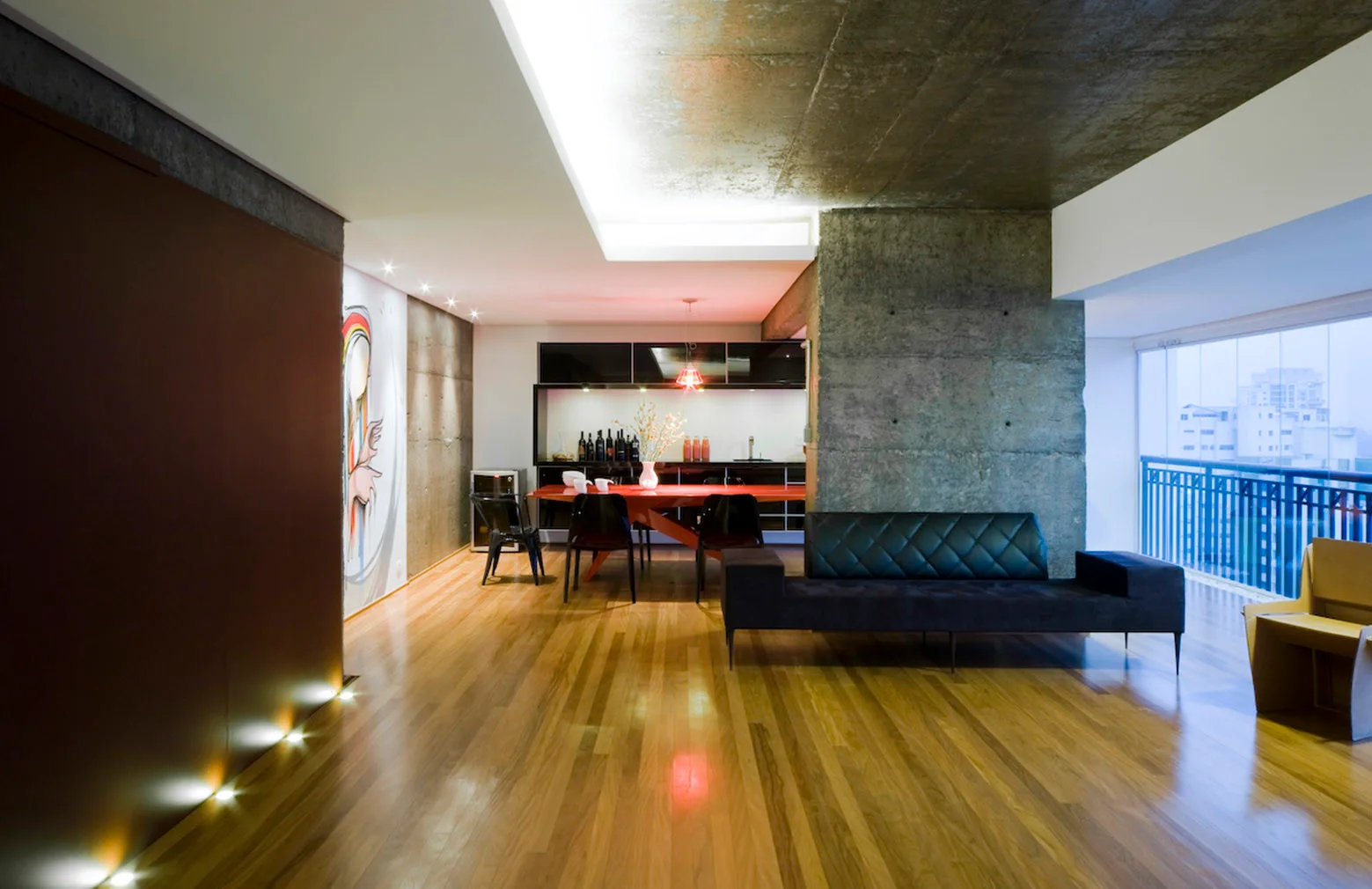An industrial open-plan living room featuring an exposed concrete ceiling, a black sofa, and a bright red dining table.