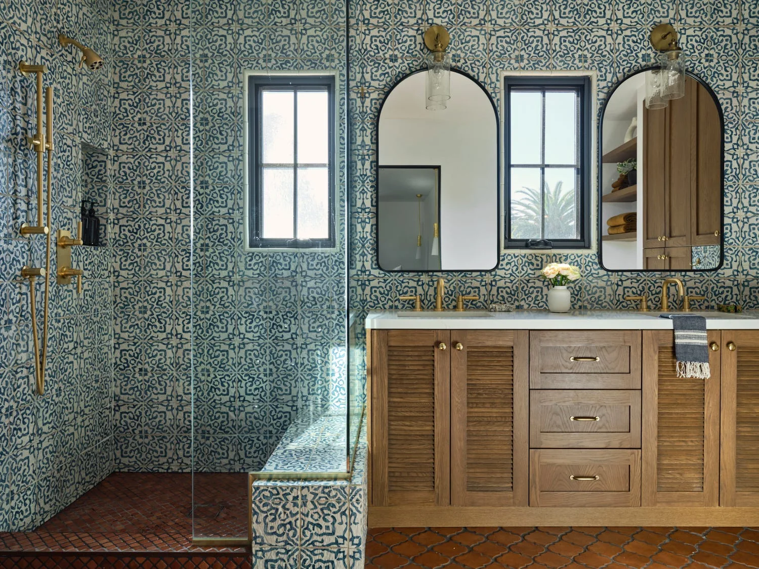 A bathroom with blue and white patterned tile walls, a wood double vanity with brass faucets, and two arched mirrors.
