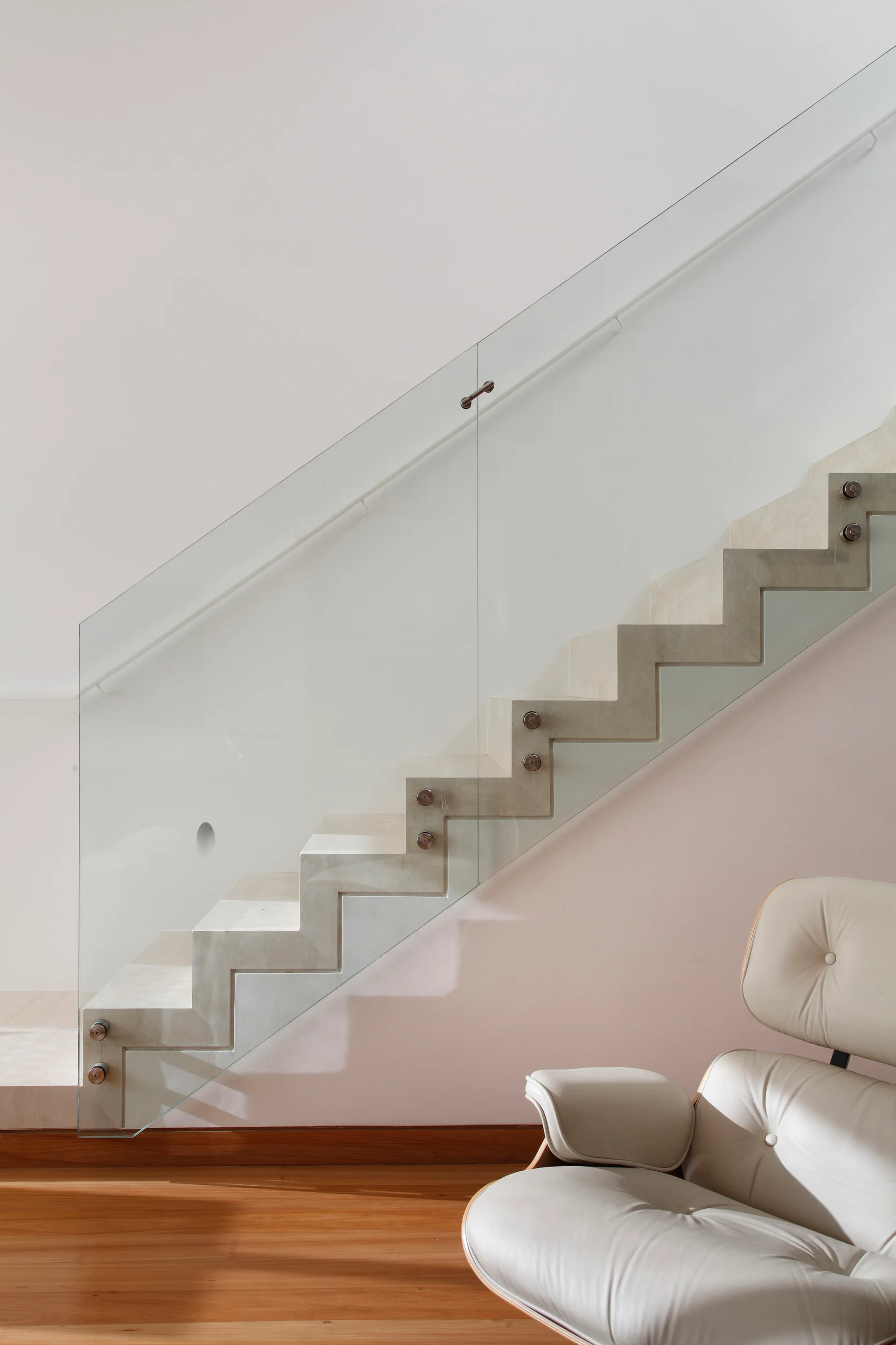 A minimalist stone zigzag staircase features a frameless glass railing next to a pale pink wall and a tufted leather lounge chair on wood floors.