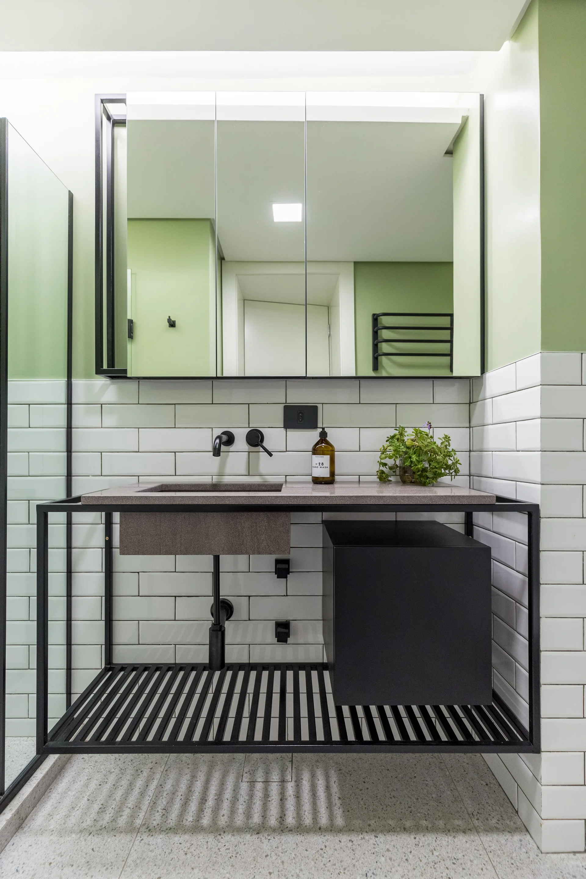 A modern bathroom vanity featuring a black metal frame, stone-like sink, white subway tile wall cladding, and a sage green upper wall.