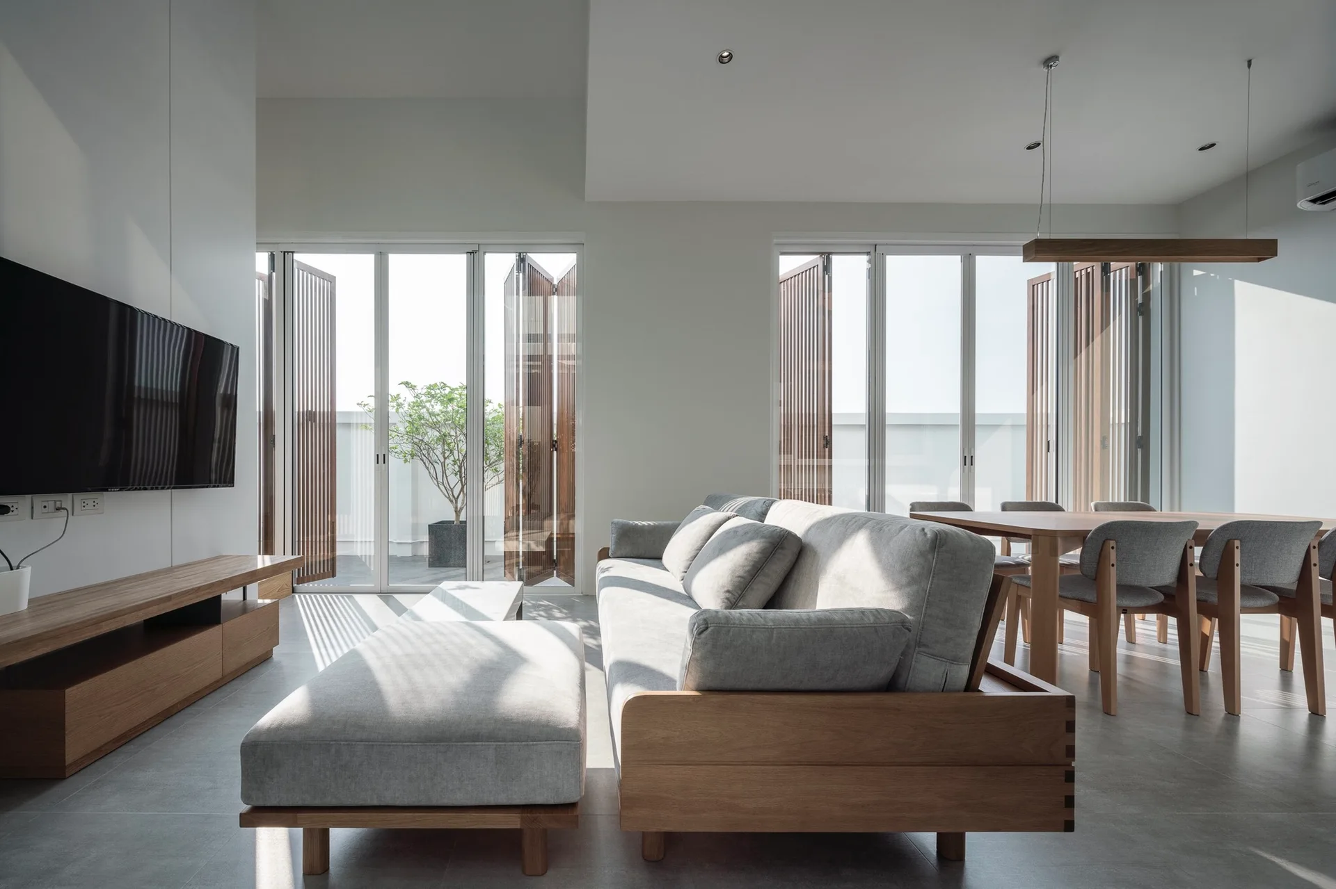 A minimalist living and dining space featuring a wood-framed grey sofa and folding glass doors with vertical slatted wood screens.