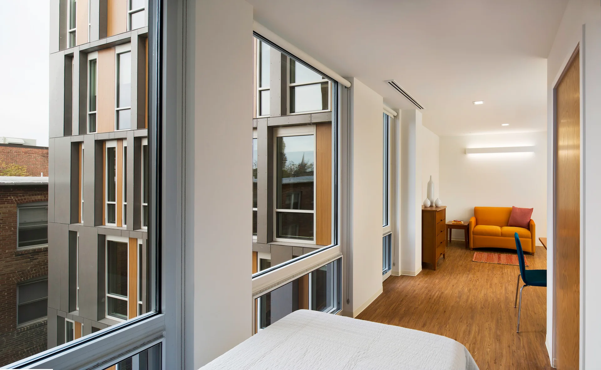 A modern apartment living room featuring an orange sofa, a blue chair, wood floors, and large windows looking out at an adjacent building facade.